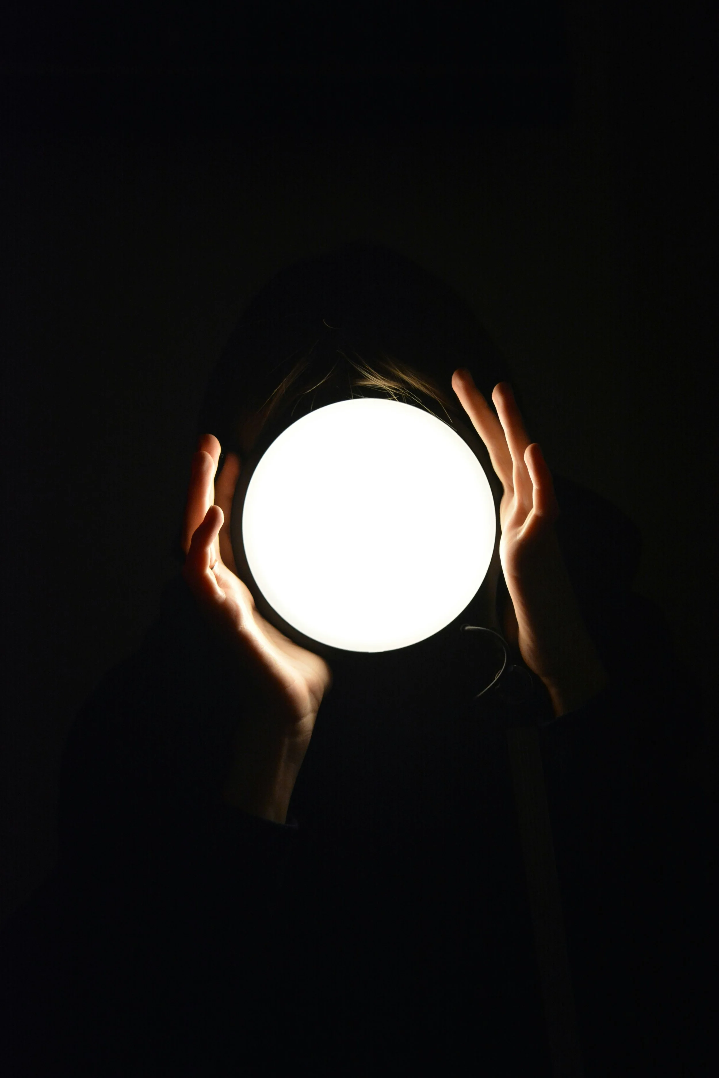 Person holding a bright, round light source in front of their face, obscuring it in darkness.