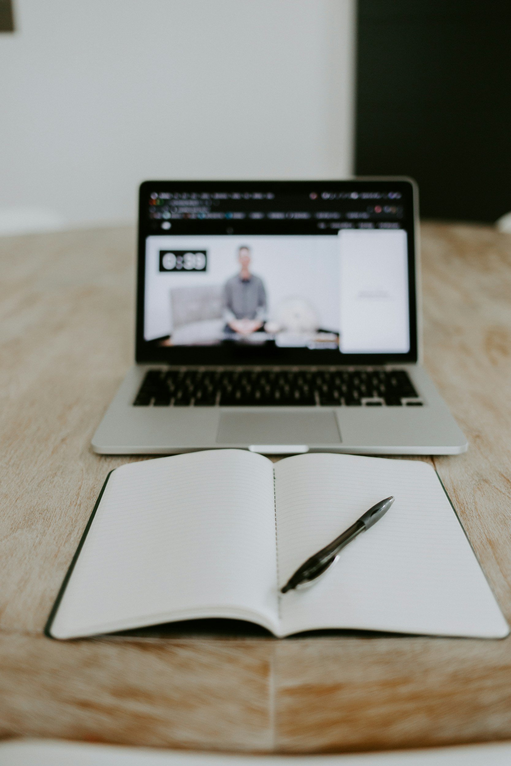 Open notebook with a pen on a wooden desk, in front of a laptop showing a blurred video conference with a person.