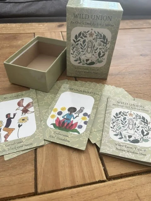 A tarot card deck named 'Wild Union' with oracle cards, featuring illustrations of a person, a bird, and a mandala, on a wooden surface. The deck box has a lotus and floral design.”}