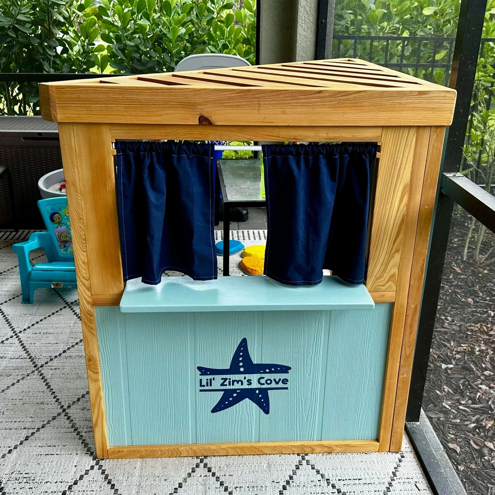 OUTDOOR CREATIVE PLAY CORNER, St. Pete, FL | Two-sided toddler play station with a slatted pergola-style roof for charm and shade. This side features a peek-through window and counter framed with washable outdoor curtains  - perfect for puppet shows,