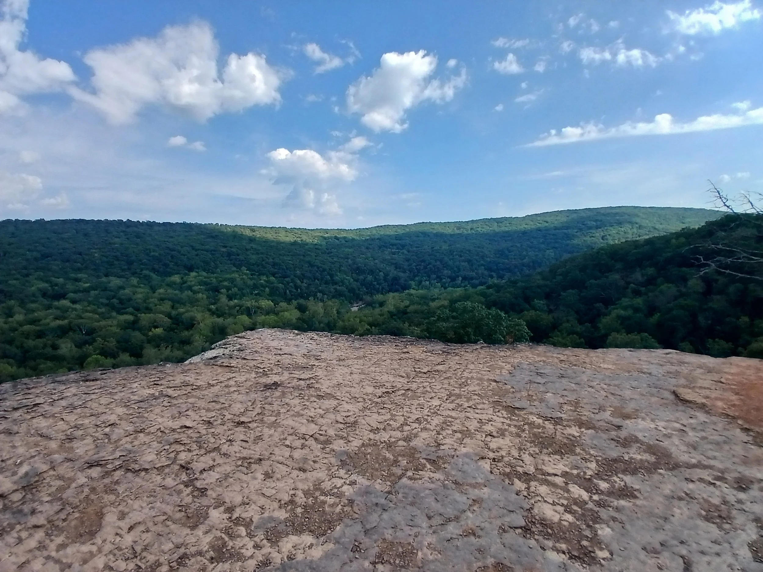 A scenic view of green forested hills under a blue sky with scattered white clouds, viewed from a rocky ledge in the foreground.