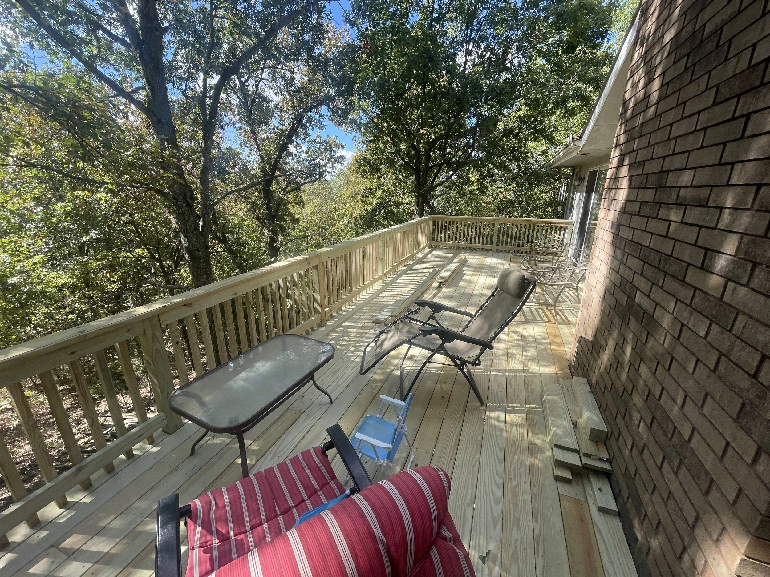 A wooden balcony with outdoor chairs and a glass table, overlooking a lush green forest with tall trees and clear blue sky.