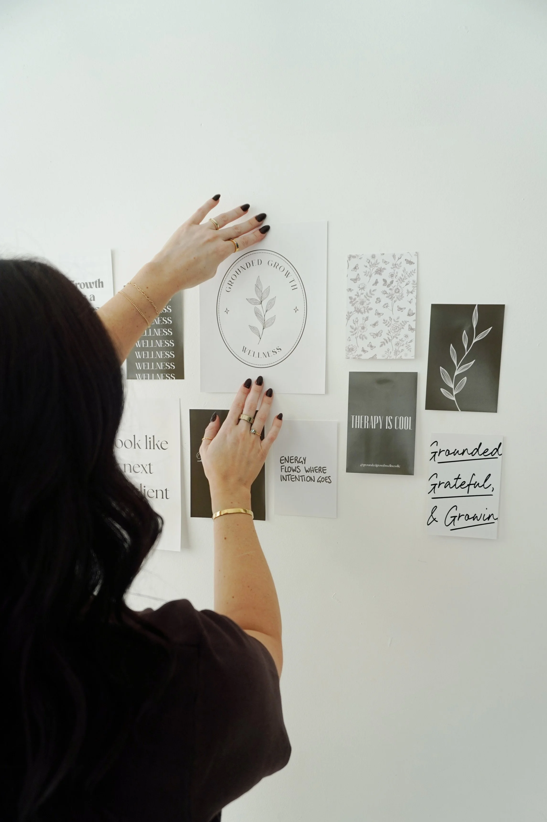 A woman with dark hair and painted black nails is placing or adjusting papers on a white wall. The papers have black and white designs and text, including a circle with a leaf inside that reads 'Grounded Growth Wellness'.