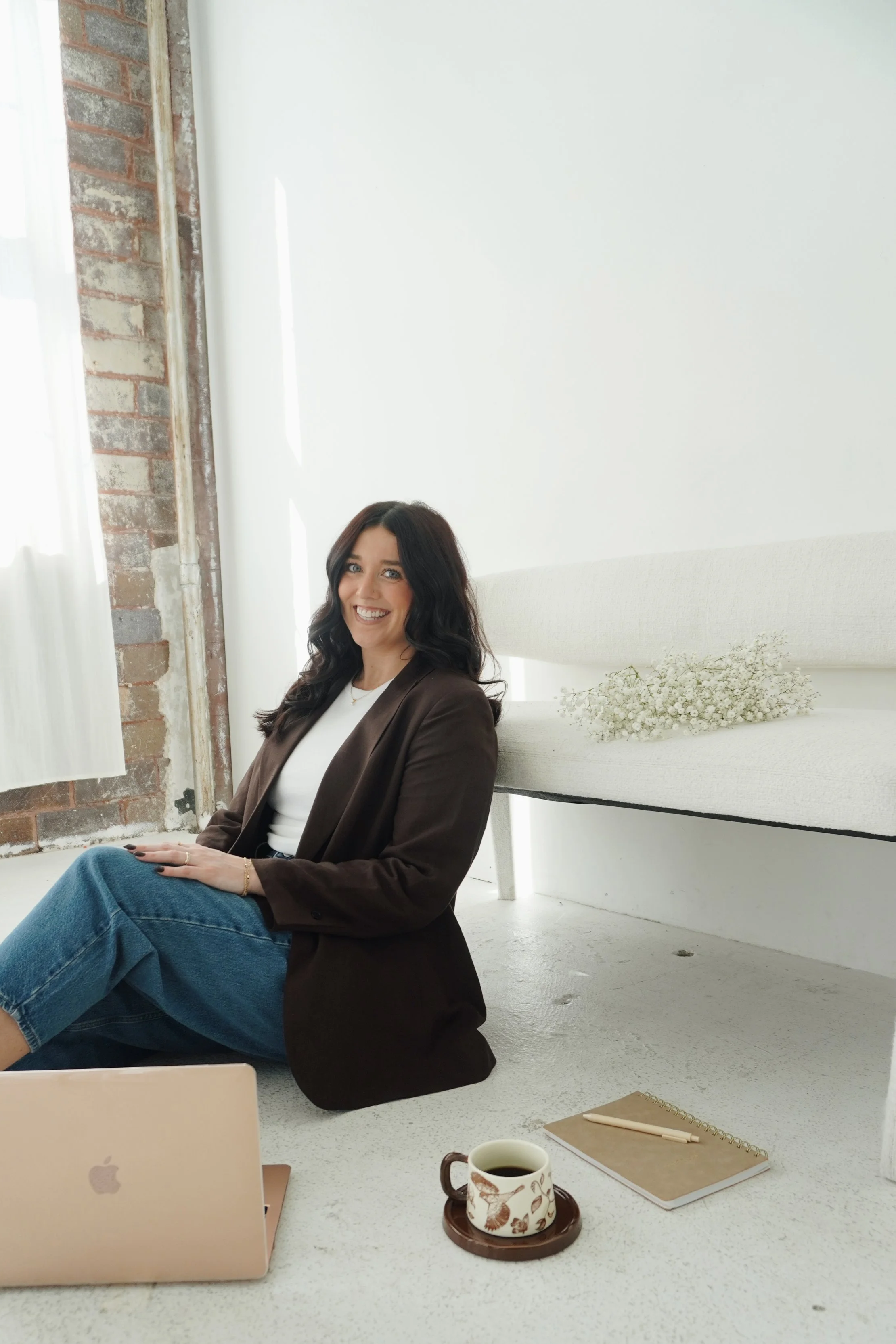 A woman with dark wavy hair sitting on the floor in a bright, modern room with a brick wall and white wall. She is smiling, wearing a brown blazer and jeans. On the floor in front of her is a laptop, a coffee mug, a notebook, and a pen.