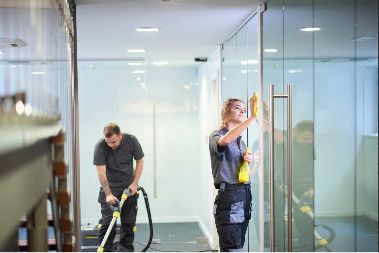 Two cleaning workers from Columbus Cleaning Service in an office corridor cleaning glass doors wearing cleaning cloths and holding spray bottles.