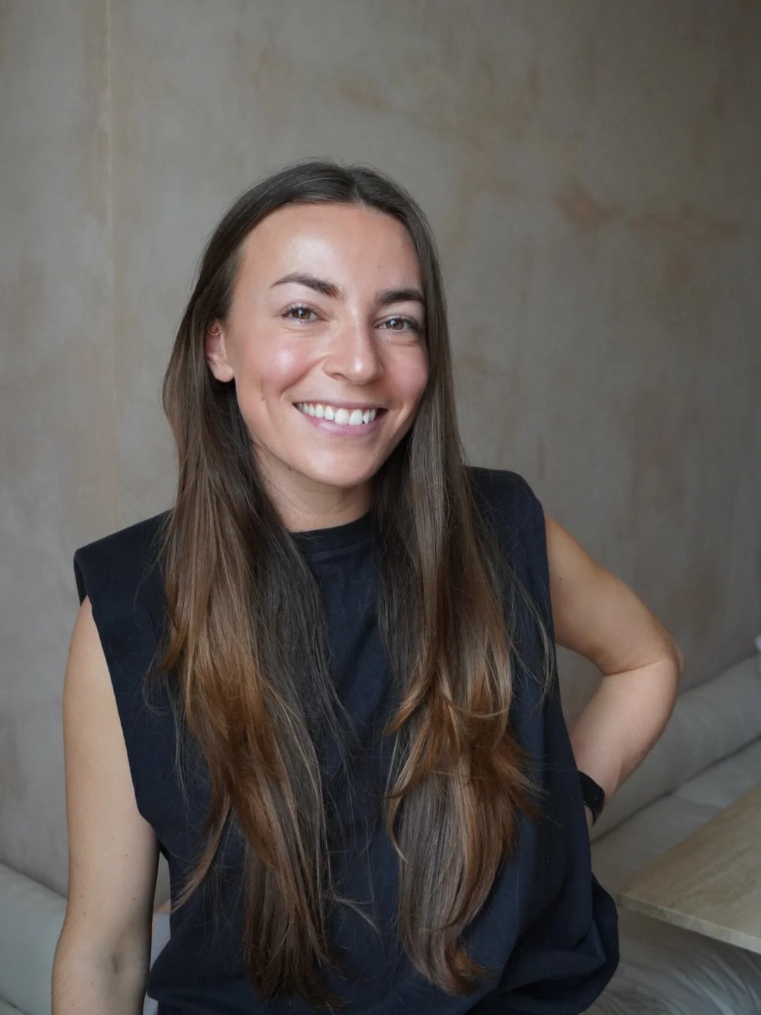 Smiling woman Ruby Deyong Andrews with long brown hair wearing a black sleeveless top, sitting in a room with beige walls and a beige sofa.
