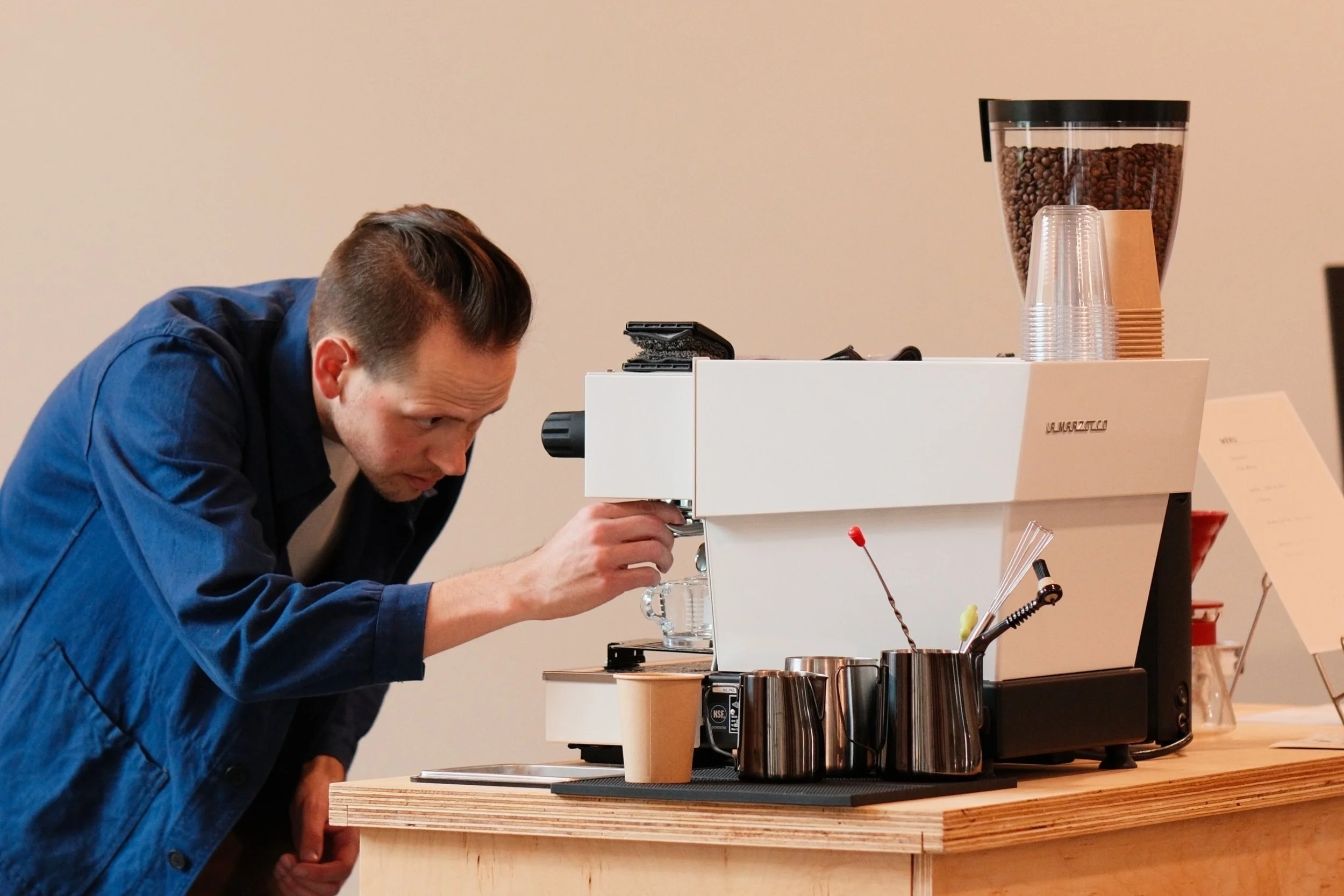 A man in a blue jacket leaning over a coffee machine, preparing coffee, with a row of stainless steel milk frothing pitchers on a wooden counter.