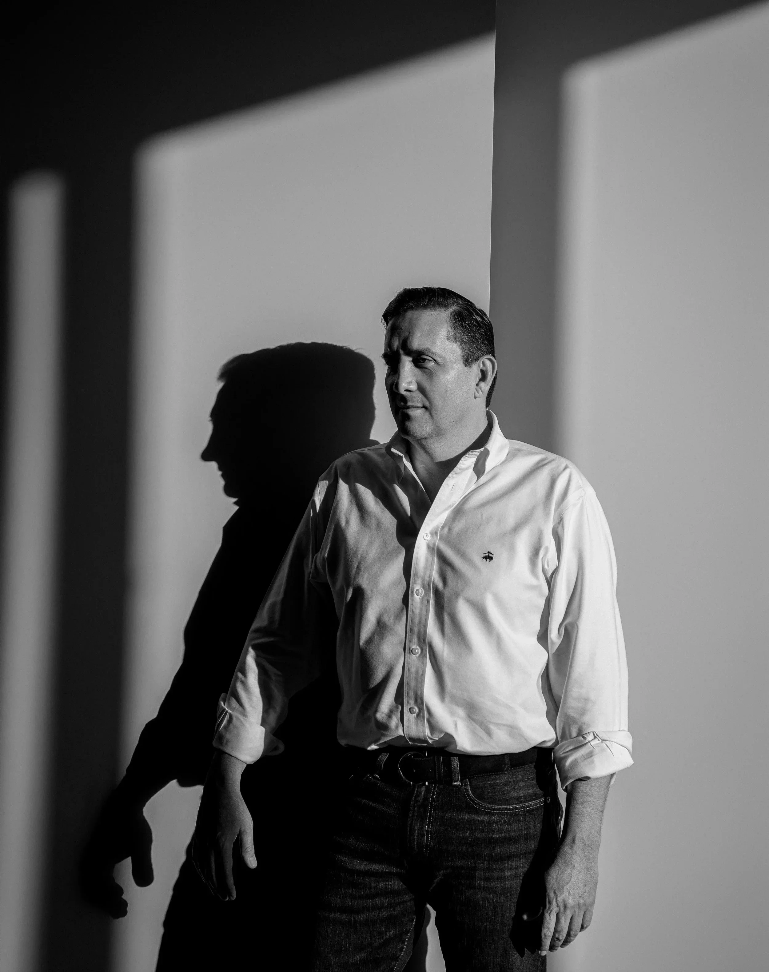 Retrato en blanco y negro de un hombre con camisa blanca junto a una pared con luz y sombras
