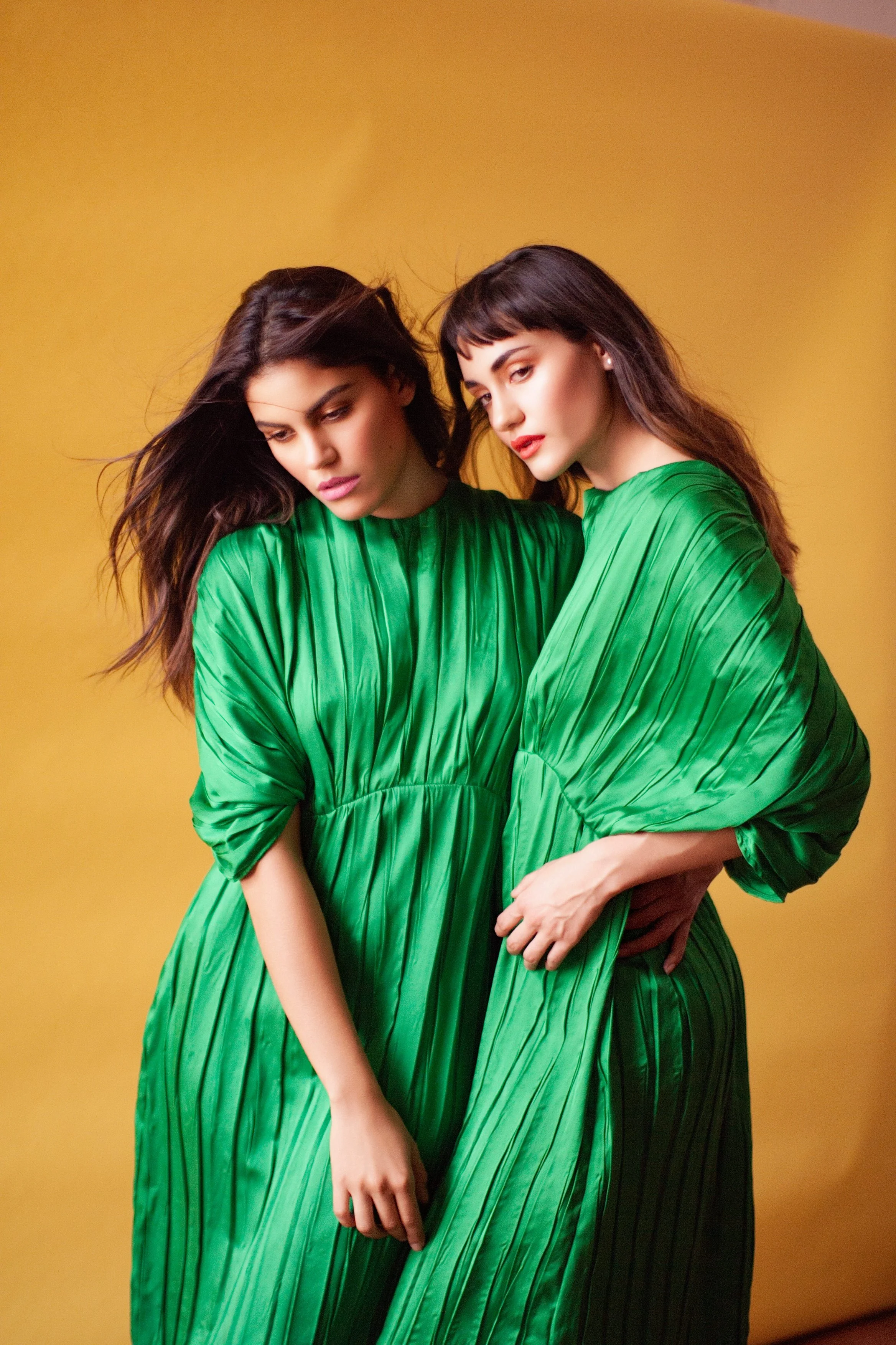 Editorial fashion portrait of two women wearing green dresses, photographed against a bold color background with balanced composition.