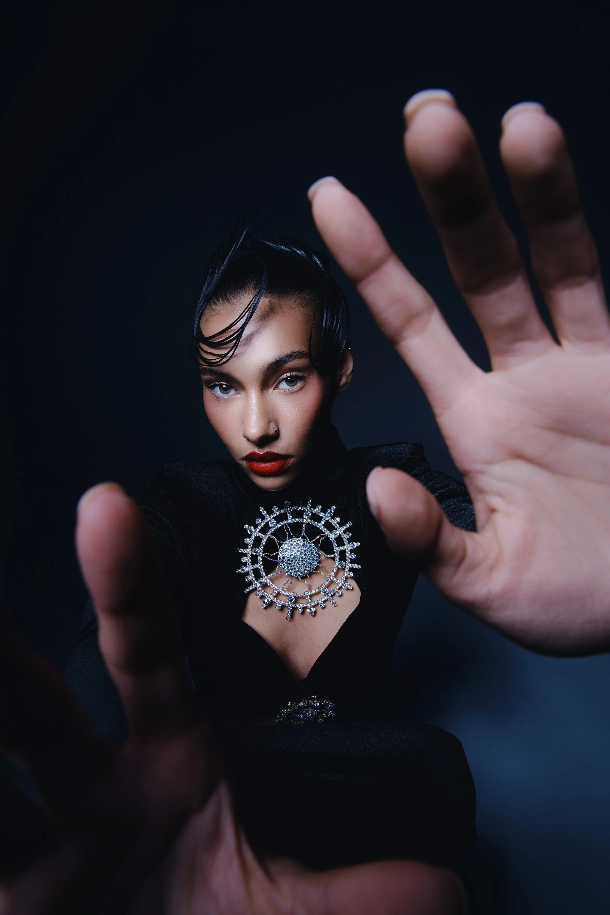 Editorial beauty portrait of a woman extending her hands toward the camera, wearing a circular statement jewelry piece, photographed with dramatic studio lighting.