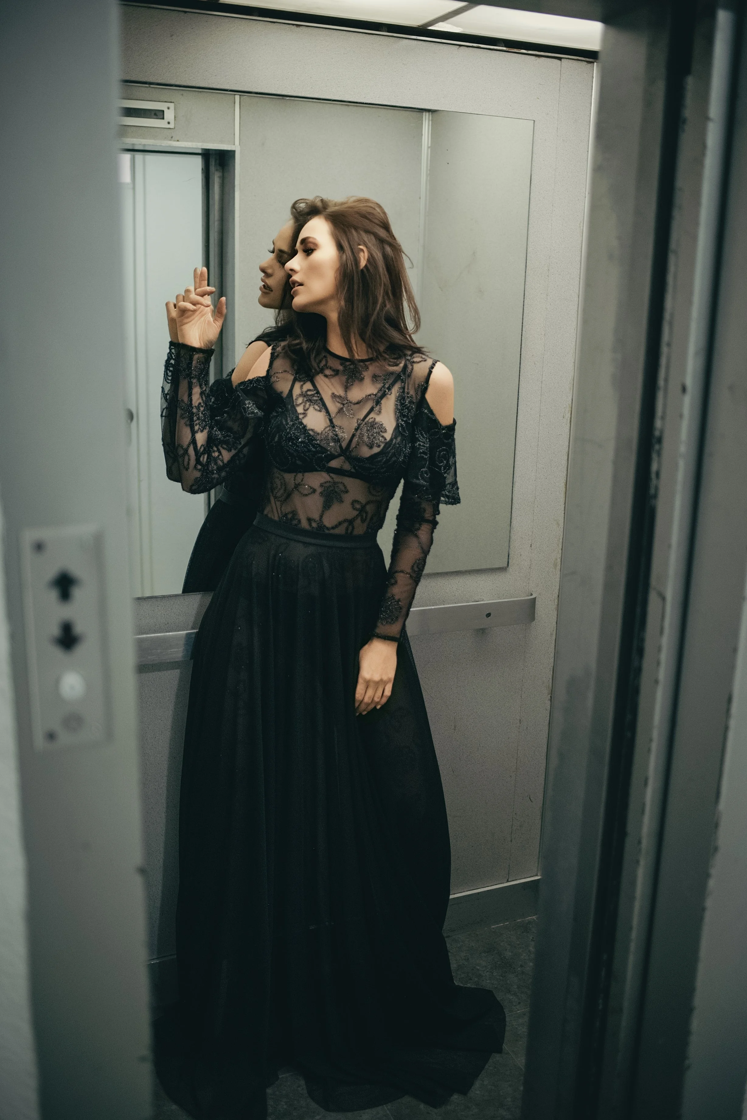 Editorial fashion portrait of a woman standing inside an elevator, wearing a black lace outfit, captured in a candid pose under soft interior lighting.