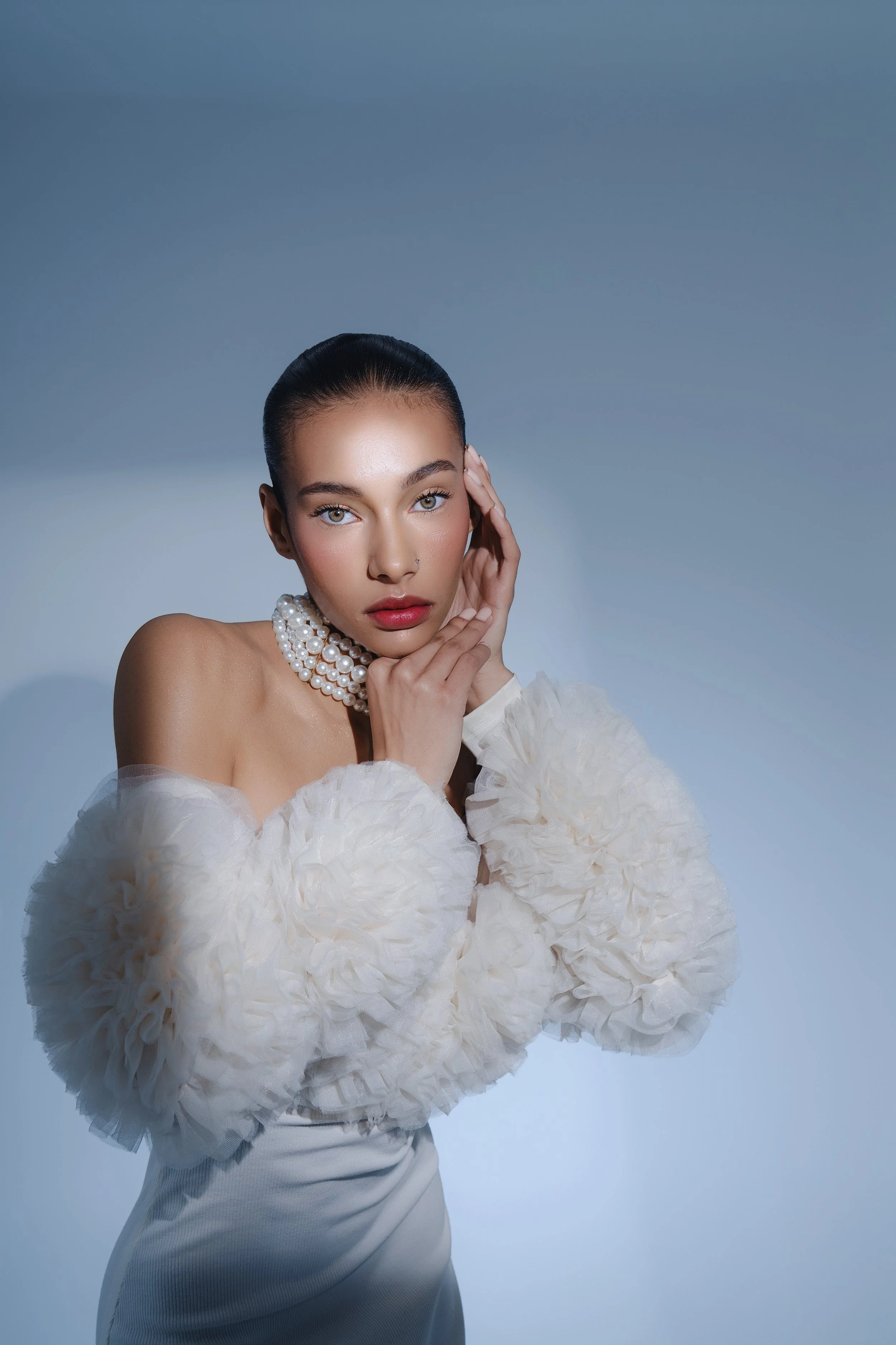 Retrato de una mujer con vestido blanco y collar de perlas, mano en el rostro, sobre fondo azul claro