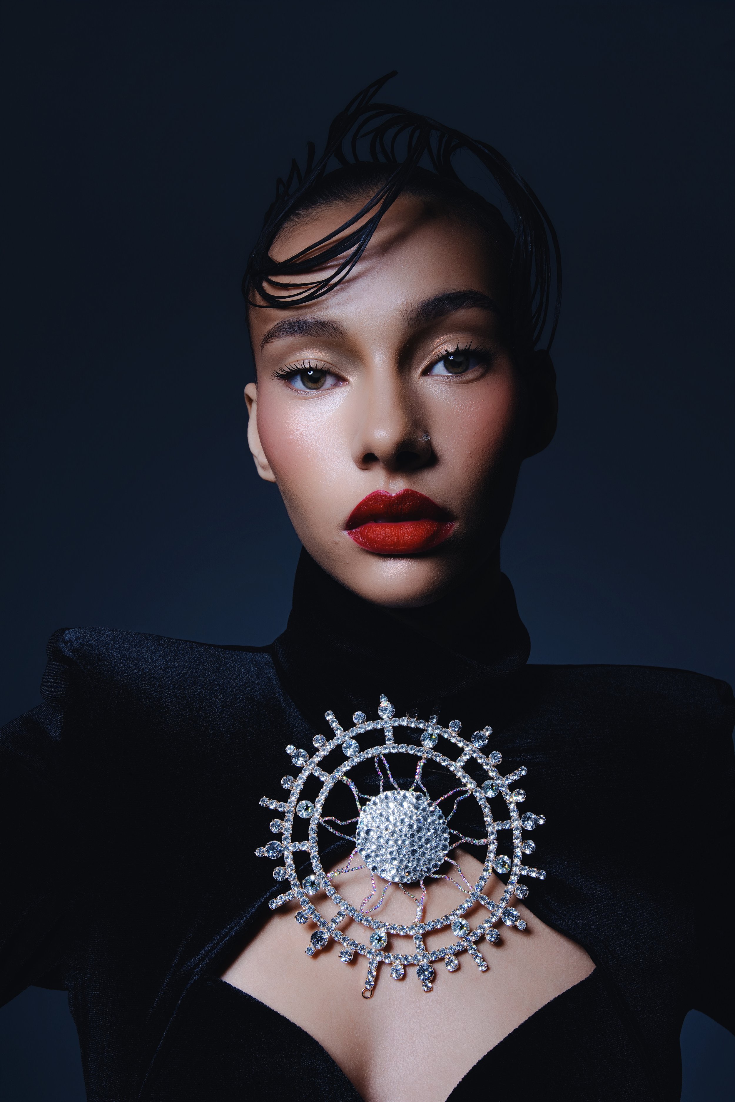 Frontal editorial beauty portrait of a woman wearing a sculptural jewelry piece, photographed against a dark background with controlled light.