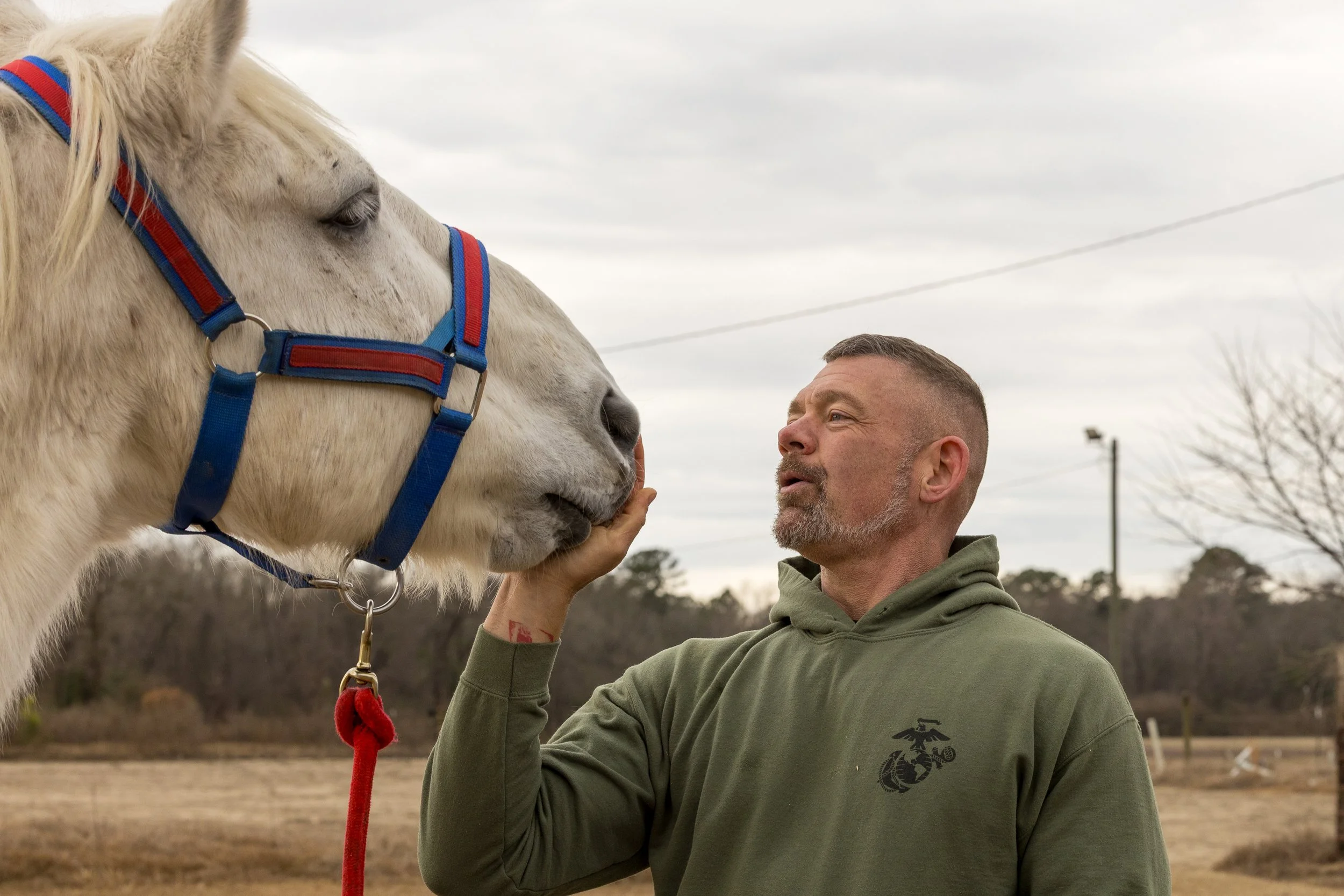 A man holding a white horse's face close to his own, outdoors on a cloudy day.