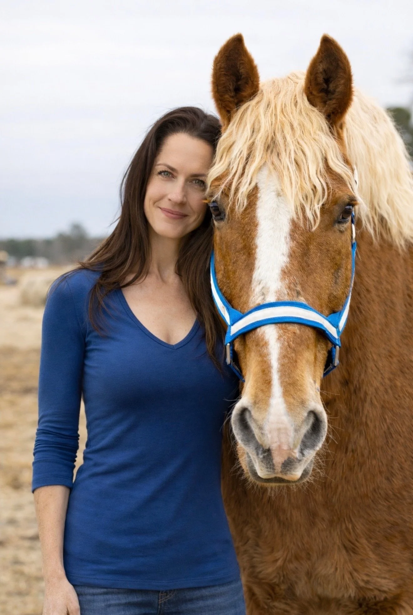 A woman with long dark hair wearing a blue top standing next to a brown horse with a blonde mane, both facing forward outdoors.