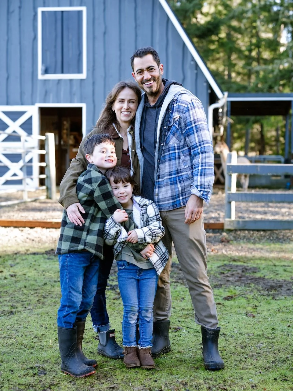 Smiling family photo in front of a blue barn. Daria hugs his wife while son and daughter stand in front.