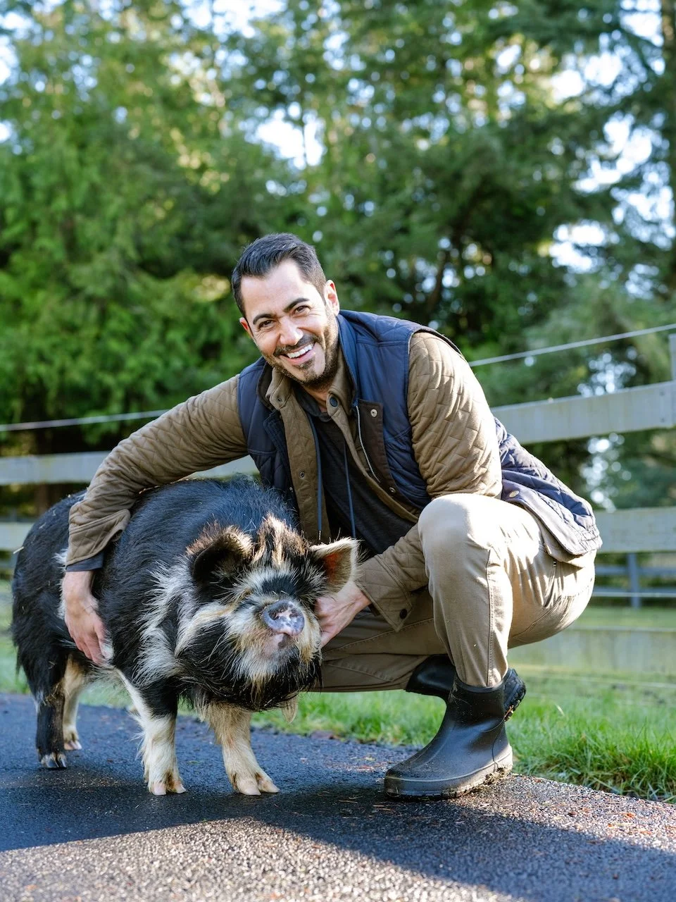 Daria crouched down smiling and hugging a large black and white pig. He is wearing tan pants, tan jacket, navy vest, and work boots .