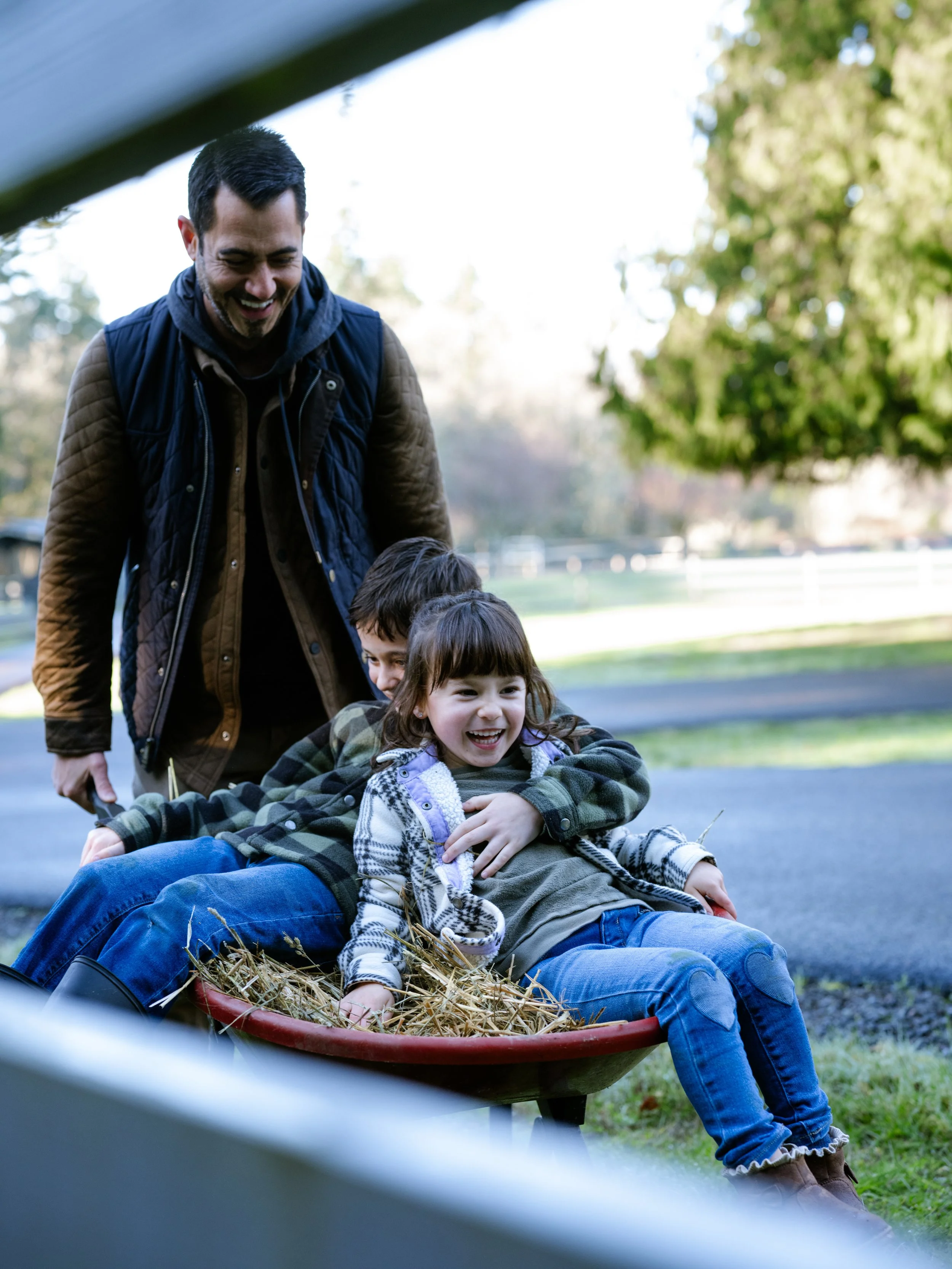 Daria pushes his two small children in a red, hay-filled wheelbarrow. All three smiling.