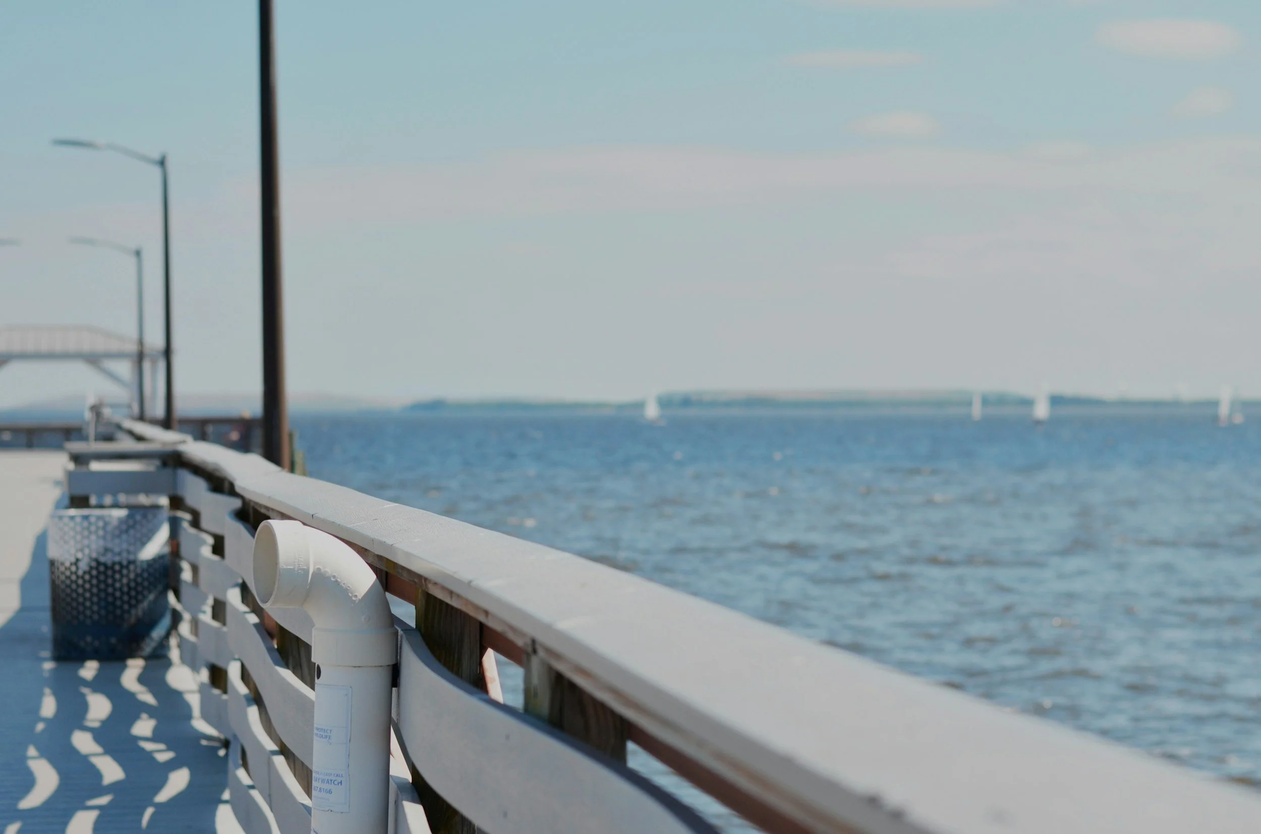 View of a waterfront walkway with a white railing, lampposts, and sailboats on the water under a blue sky.