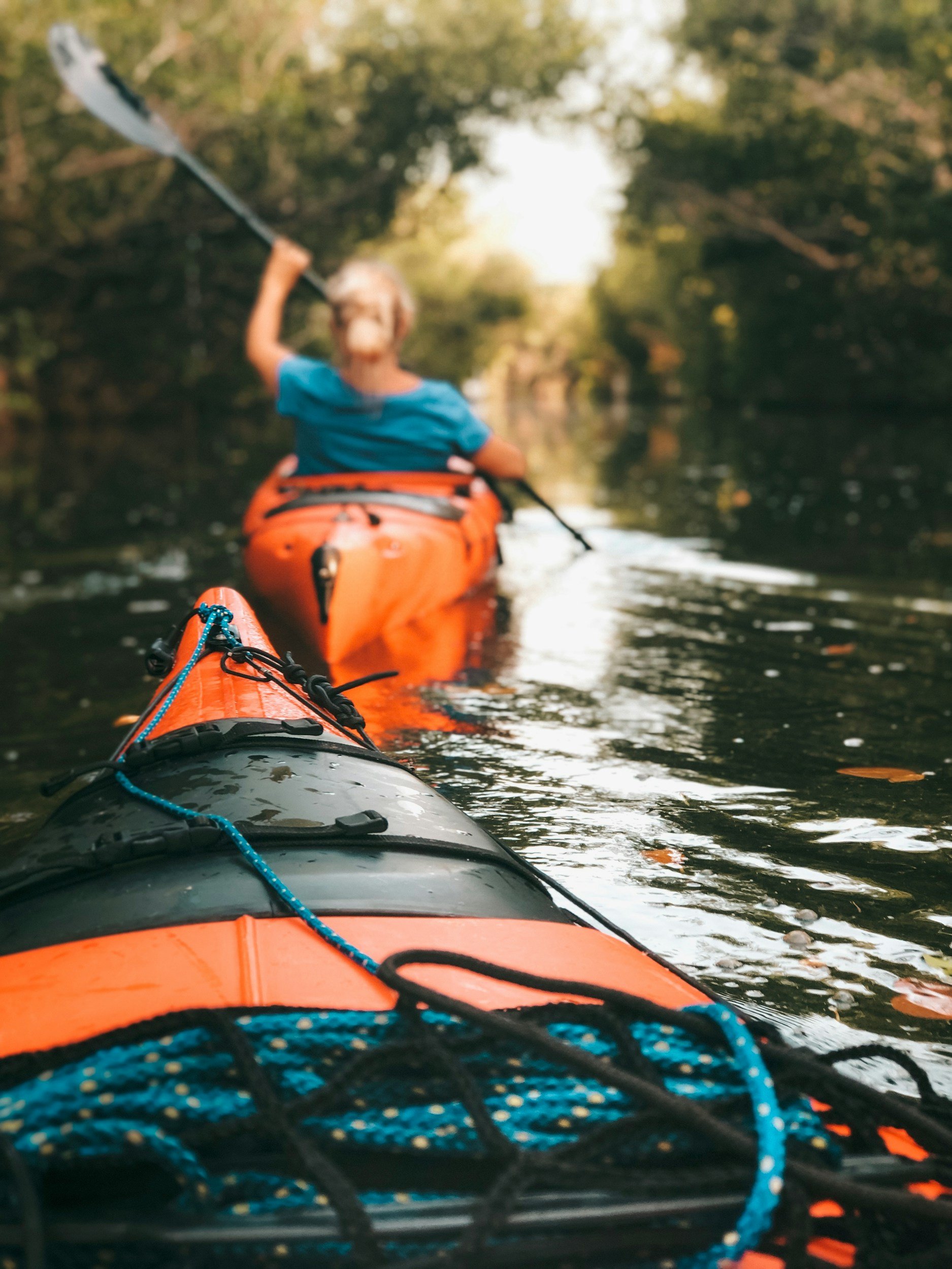 View from the front of an orange kayak on a river, with a blue and black rope securement, and a person kayaking ahead, wearing a blue shirt, paddling through a green, tree-lined waterway.