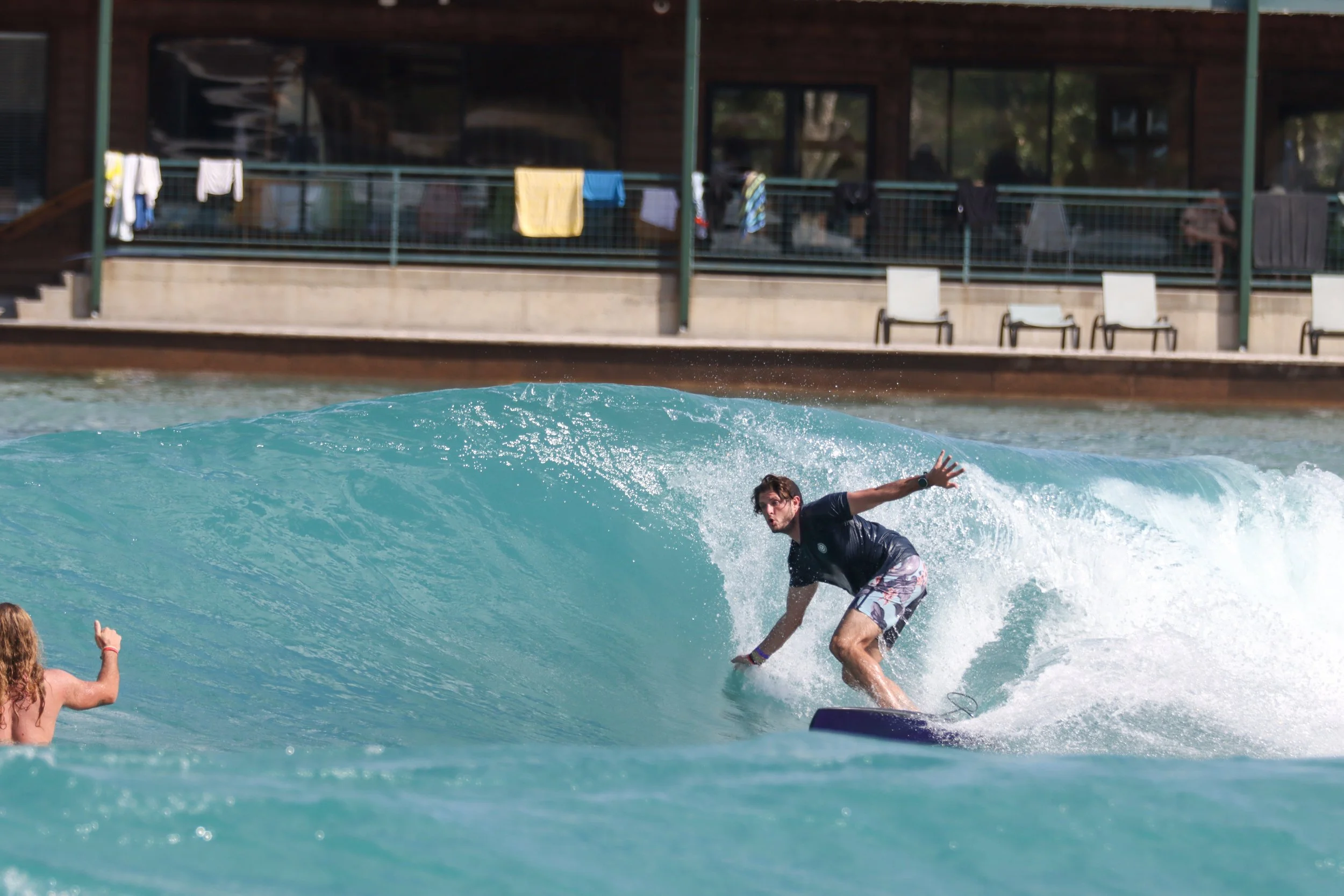 A man surfing on a wave at a beach. There is a woman in the water with her arm raised. In the background, a building with laundry hanging on a balcony and empty chairs outdoors.
