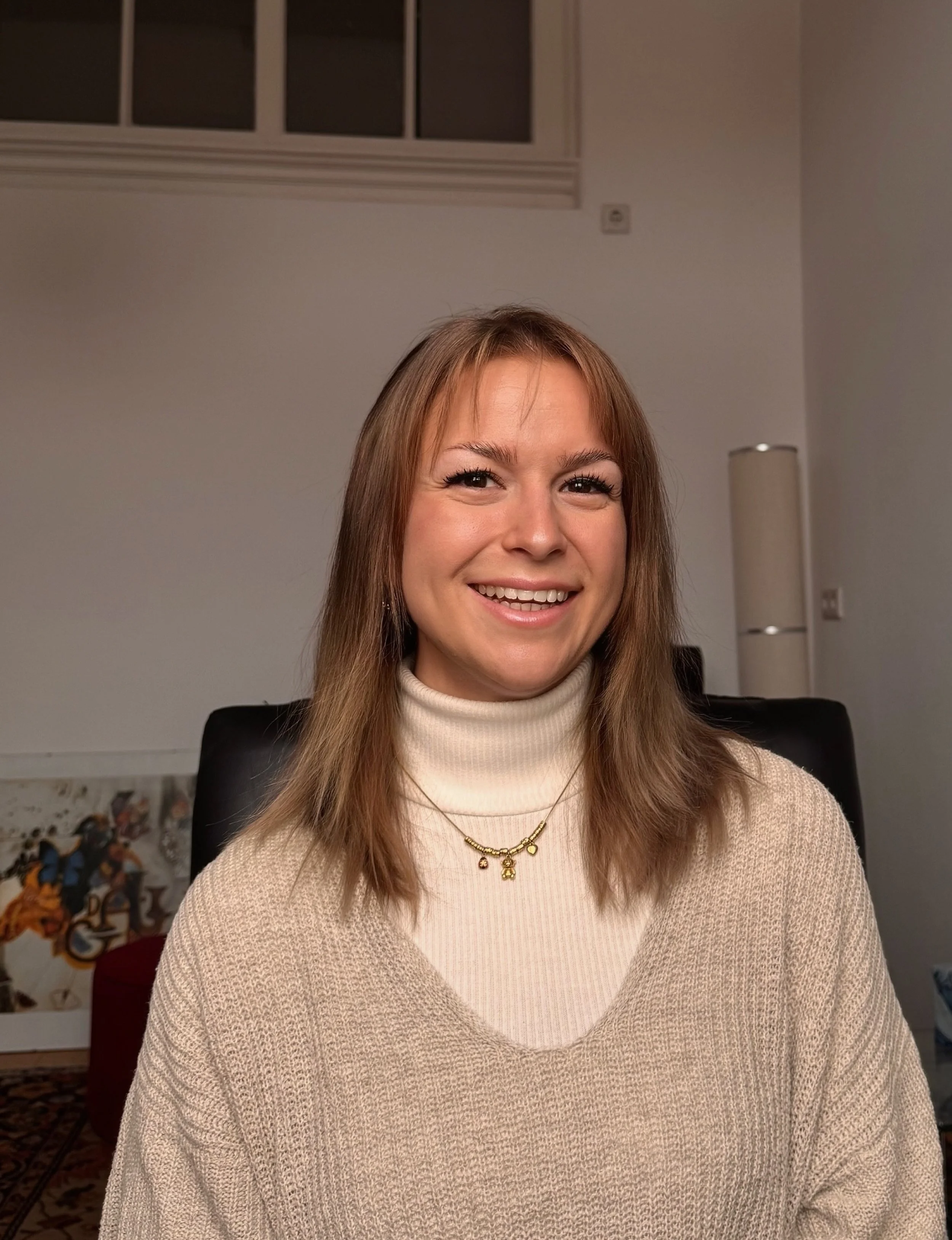 Ein Lächelndes junges Mädchen mit braunen Haaren in einem cremefarbenen Pullover sitzt in einem Raum mit weißen Wänden und einem schwarzen Stuhl.