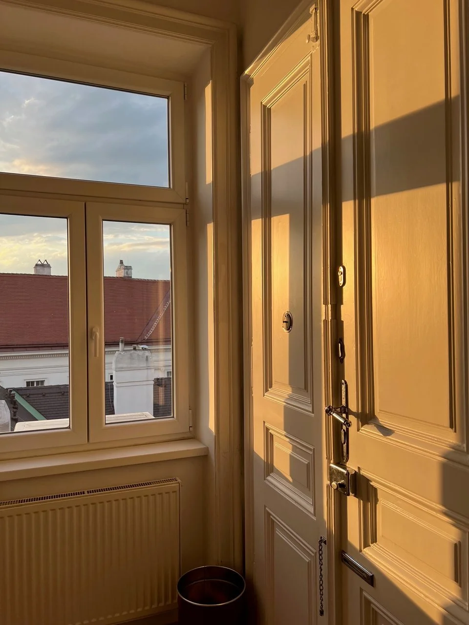 Innenansicht eines Zimmertür mit Sonnenlicht, Fenster mit Blick auf Dach und Himmel, Heizkörper unter dem Fenster, kleiner Eimer auf dem Boden.