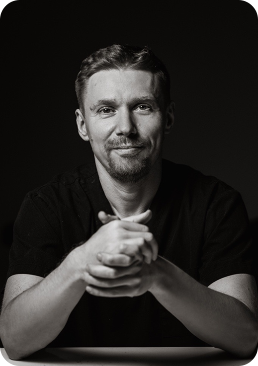 Black and white portrait of a man with short hair and a beard, sitting with hands clasped on a table, smiling slightly, in a dark background.