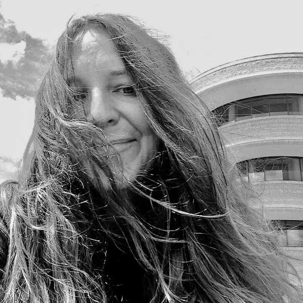 Black and white photo of a woman with long hair blowing in the wind, smiling, with a modern curved building in the background.