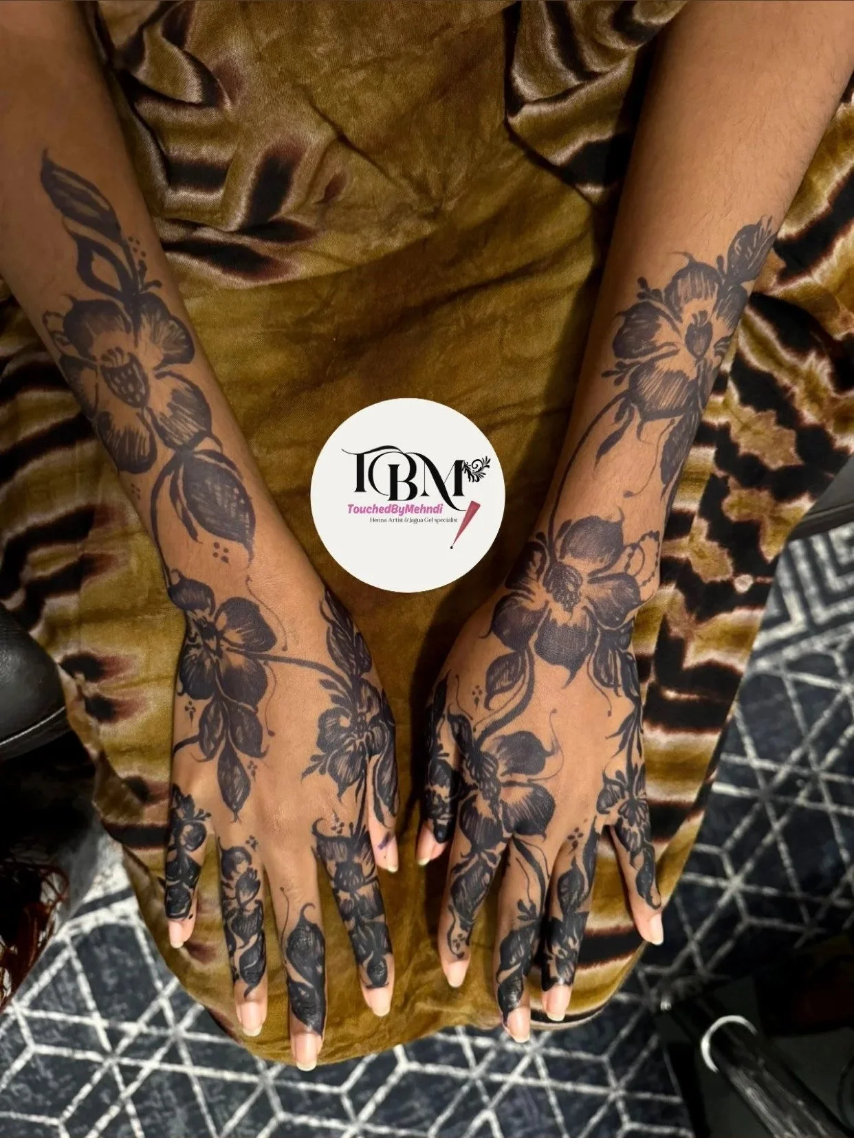 Hands with detailed black floral henna tattoos resting on a surface with a leopard print fabric, and a watermark that reads 'TouchedbyMehndi'.