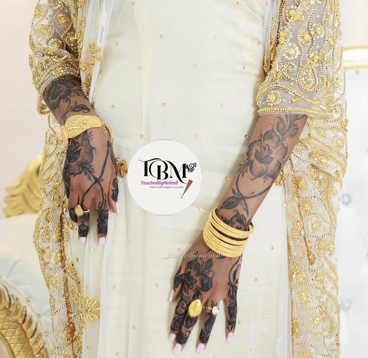 Close-up of a person dressed in an elegant, cream-colored outfit with gold embroidery. They are wearing several gold bracelets and rings. The person's hands and forearms are decorated with black floral tattoos.