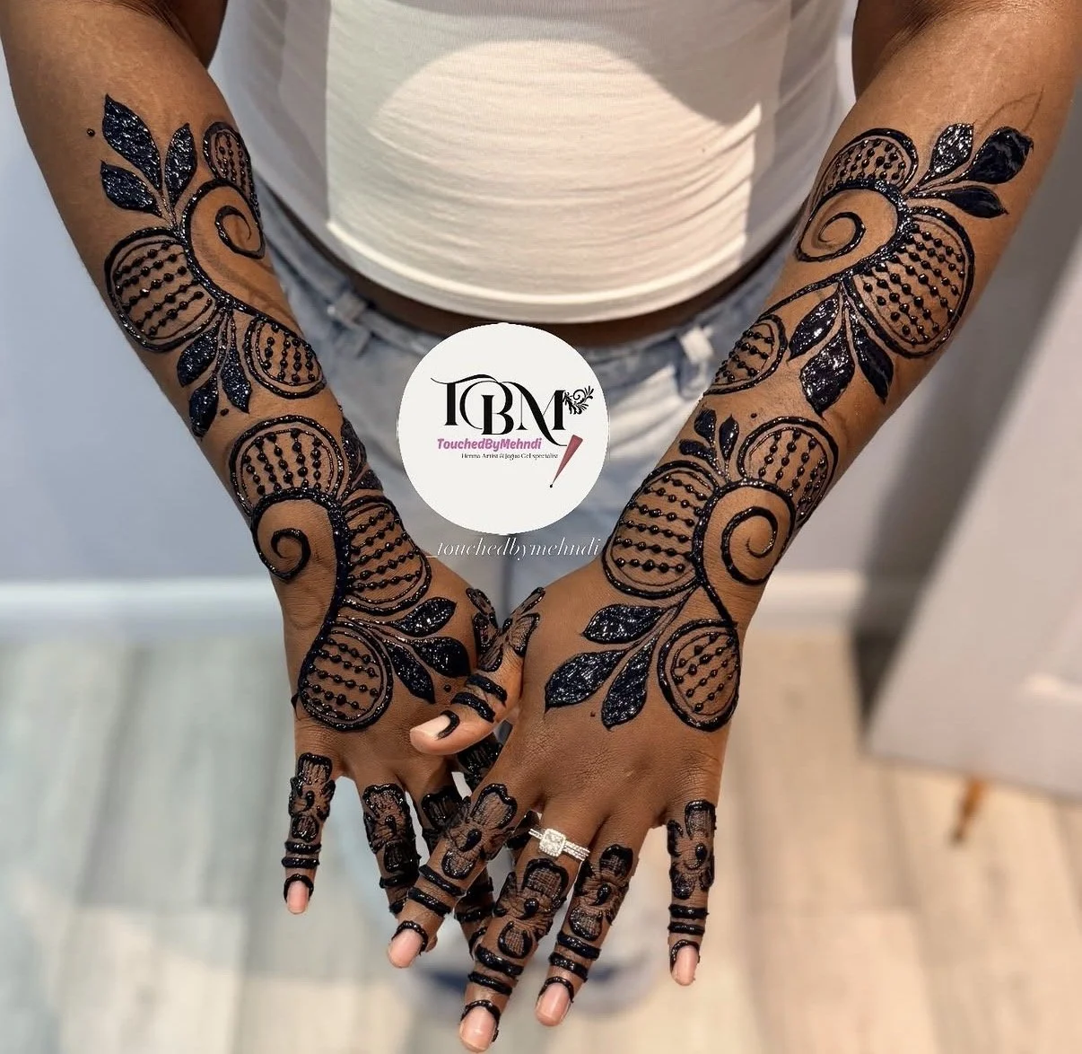 Hands with intricate black henna designs featuring floral and paisley patterns, adorned with rings, and a circular logo with text overlay.