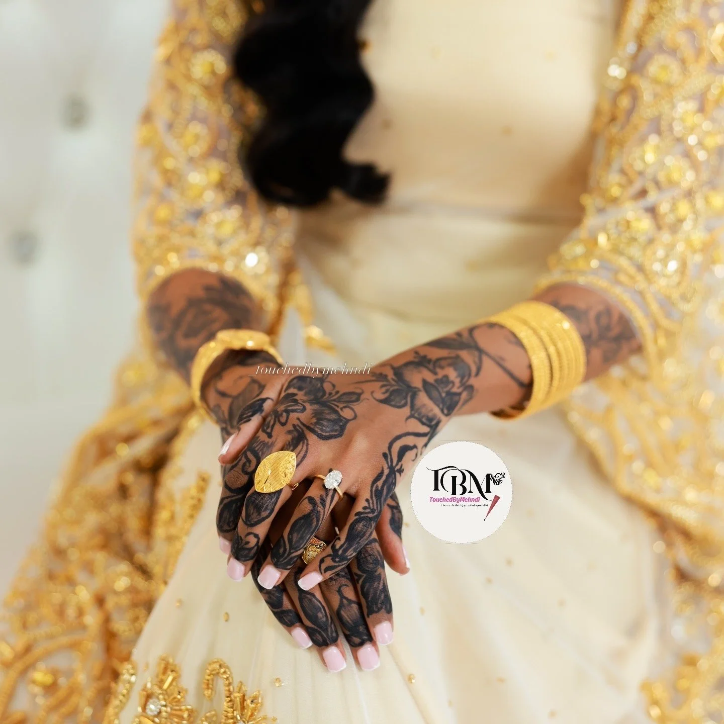 A woman wearing a gold-embroidered dress and jewelry, displaying her intricately tattooed hands and wearing rings, with her head slightly bowed.
