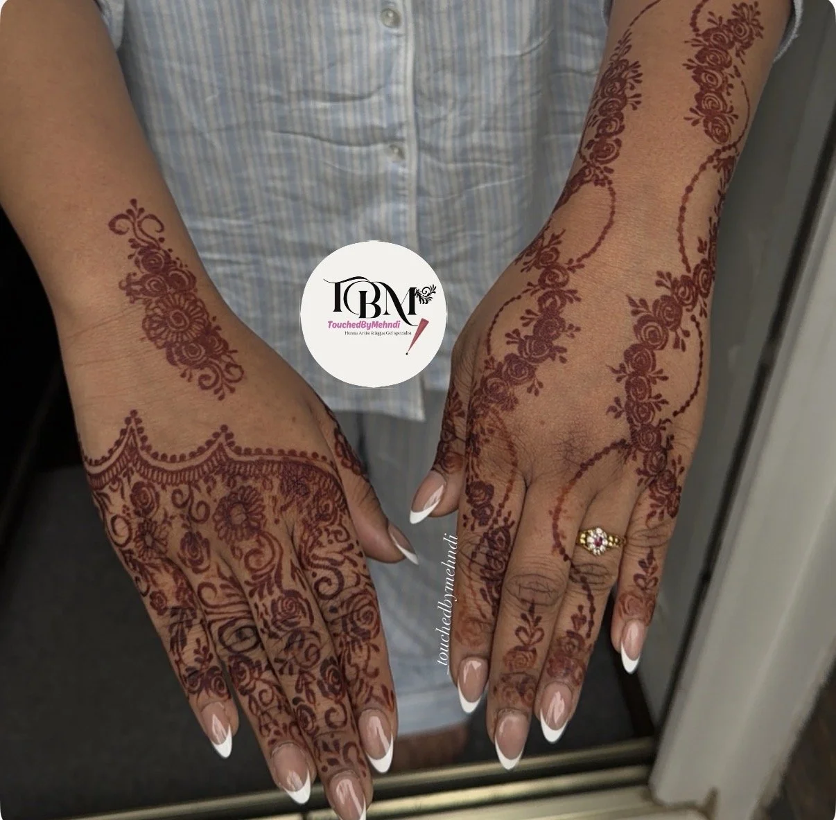 Hands with intricate henna designs on palms, fingers, and wrists, showing a gold ring on one finger, in front of a mirror reflecting the arms and henna pattern.