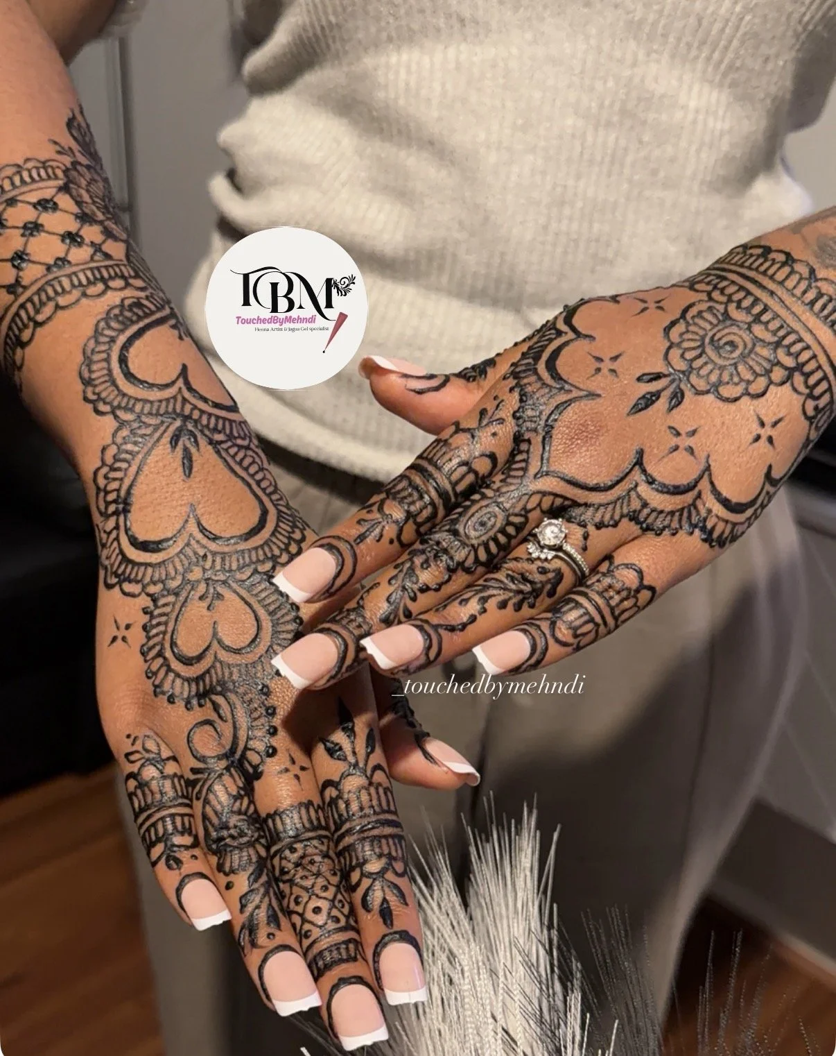 Two hands with intricate black henna designs, multiple rings, and French manicured nails, held together against gray clothing.