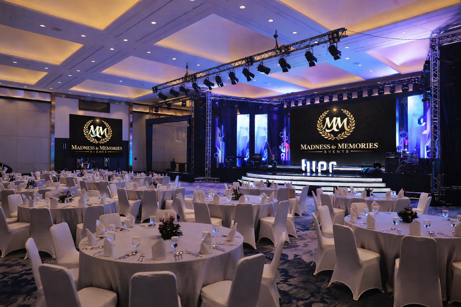 A formal event setup with round tables covered with white tablecloths, white chairs, and floral centerpieces in a large banquet hall. A stage with large screens displaying the logo and name of 'Madness to Memories Events' is in the background, illuminated with purple and blue lighting.