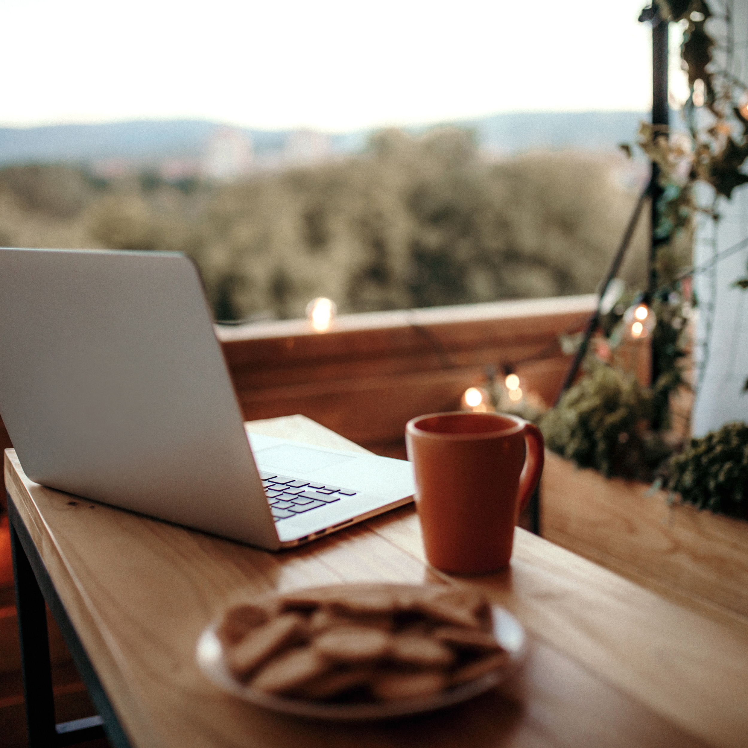 Wooden table with open laptop, orange mug, plate of cookies, on a balcony with view of trees and distant hills, string lights, and greenery.