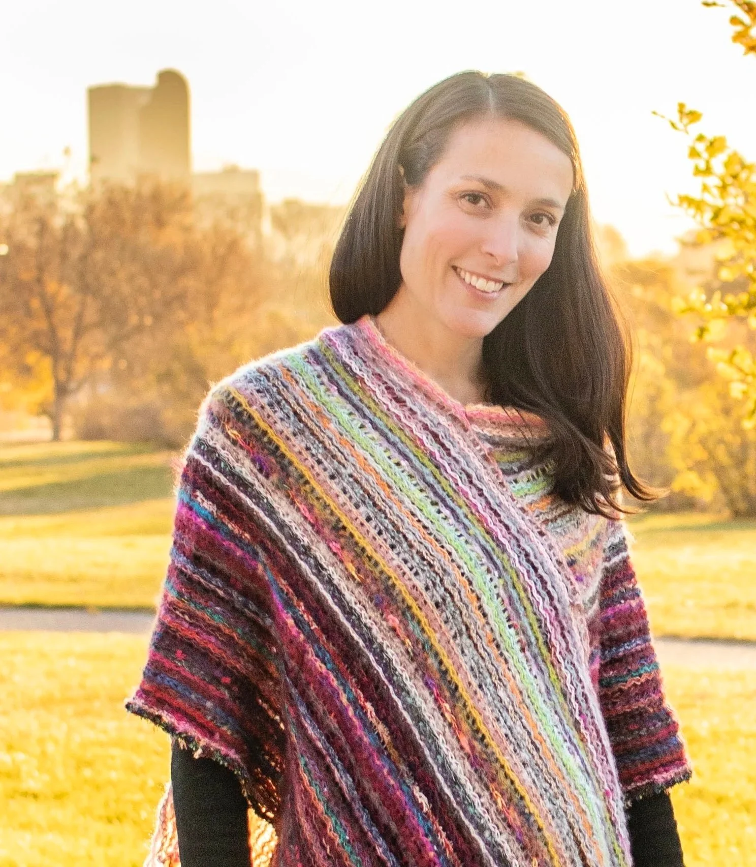 A woman with long dark hair smiling outdoors in a park during fall, wearing a colorful striped knit poncho.