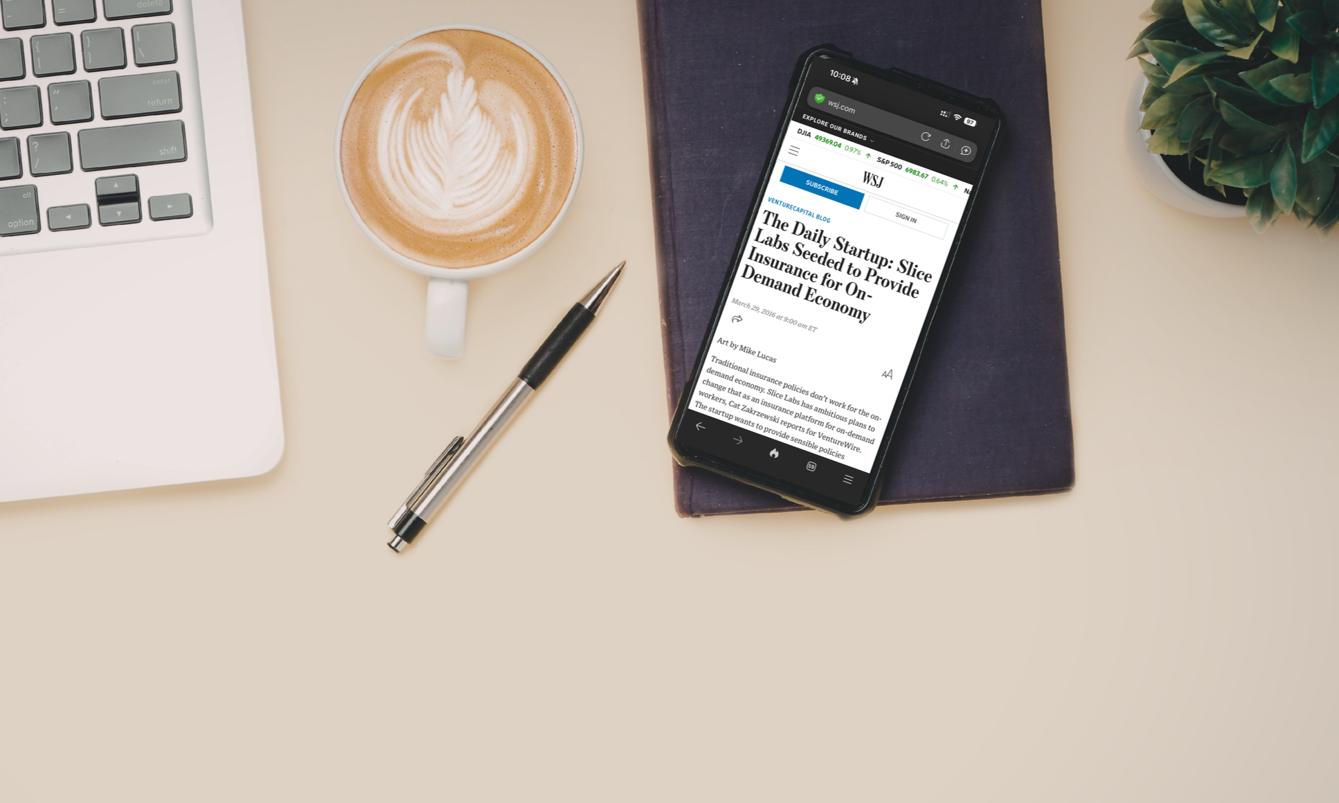 A desk with a laptop, a coffee mug with latte art, a pen, a smartphone with a news article about startups, a notebook, and a potted plant.