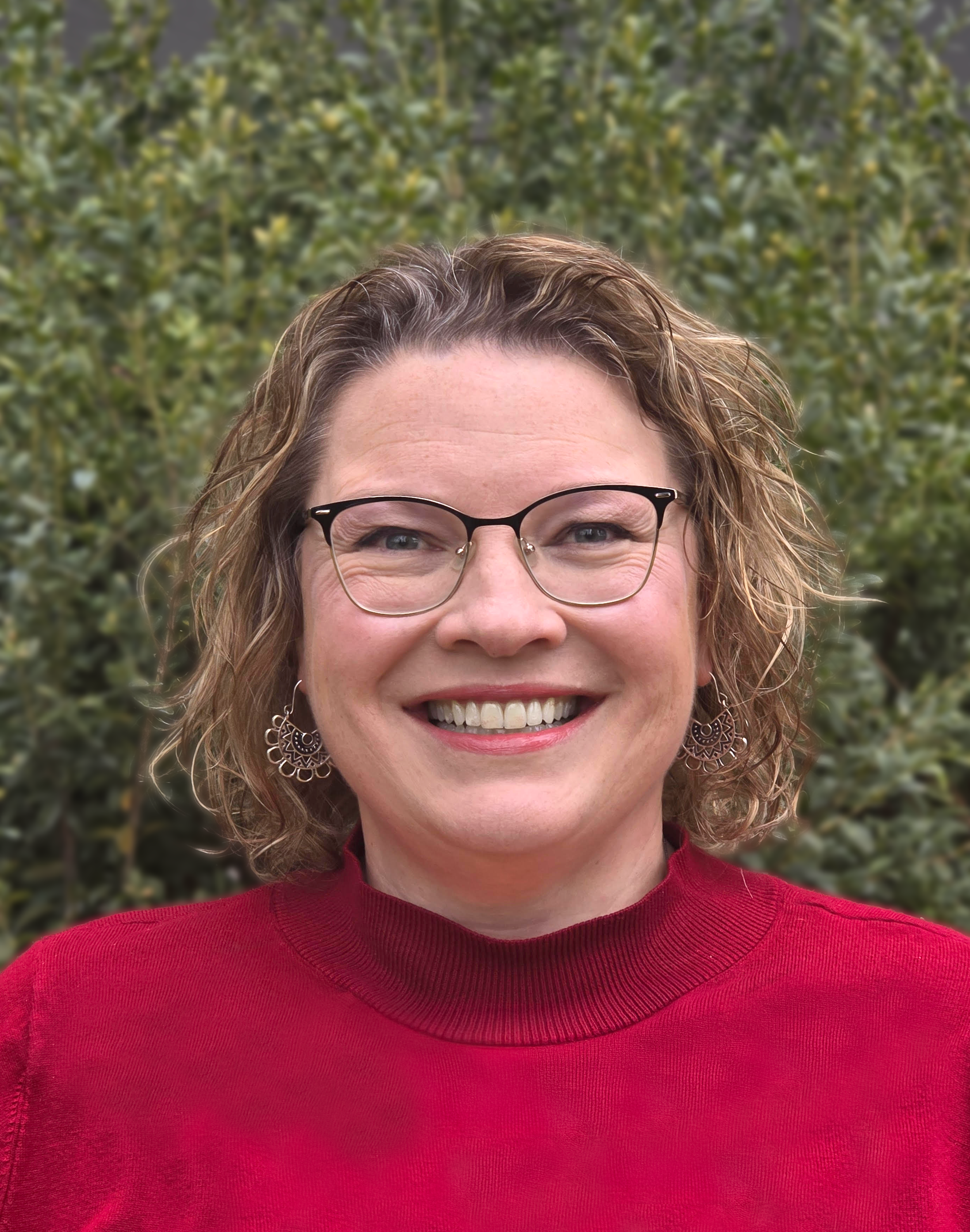 A woman with brown curly hair wearing glasses and earrings, smiling, outdoors with green foliage in the background.