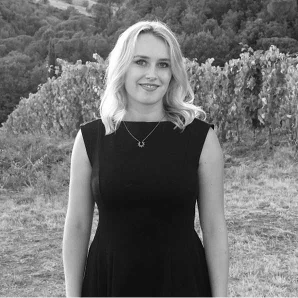 Junge Frau mit nach oben gewandter Schulter, blonden, gewellten Haaren, in einem schwarzen Kleid, steht im Freien vor einer hügeligen, bewaldeten Landschaft.