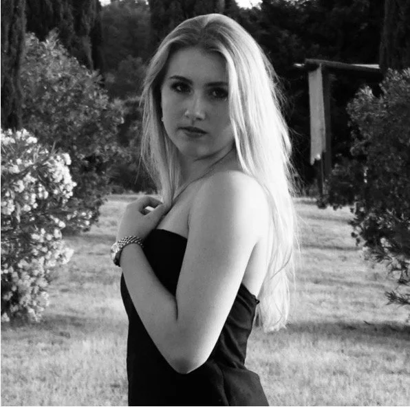 Ein junges blondes Mädchen in einem schwarzen Kleid, das im Freien steht, umgeben von Büschen und Bäumen, in Schwarz-Weiß aufgenommen.