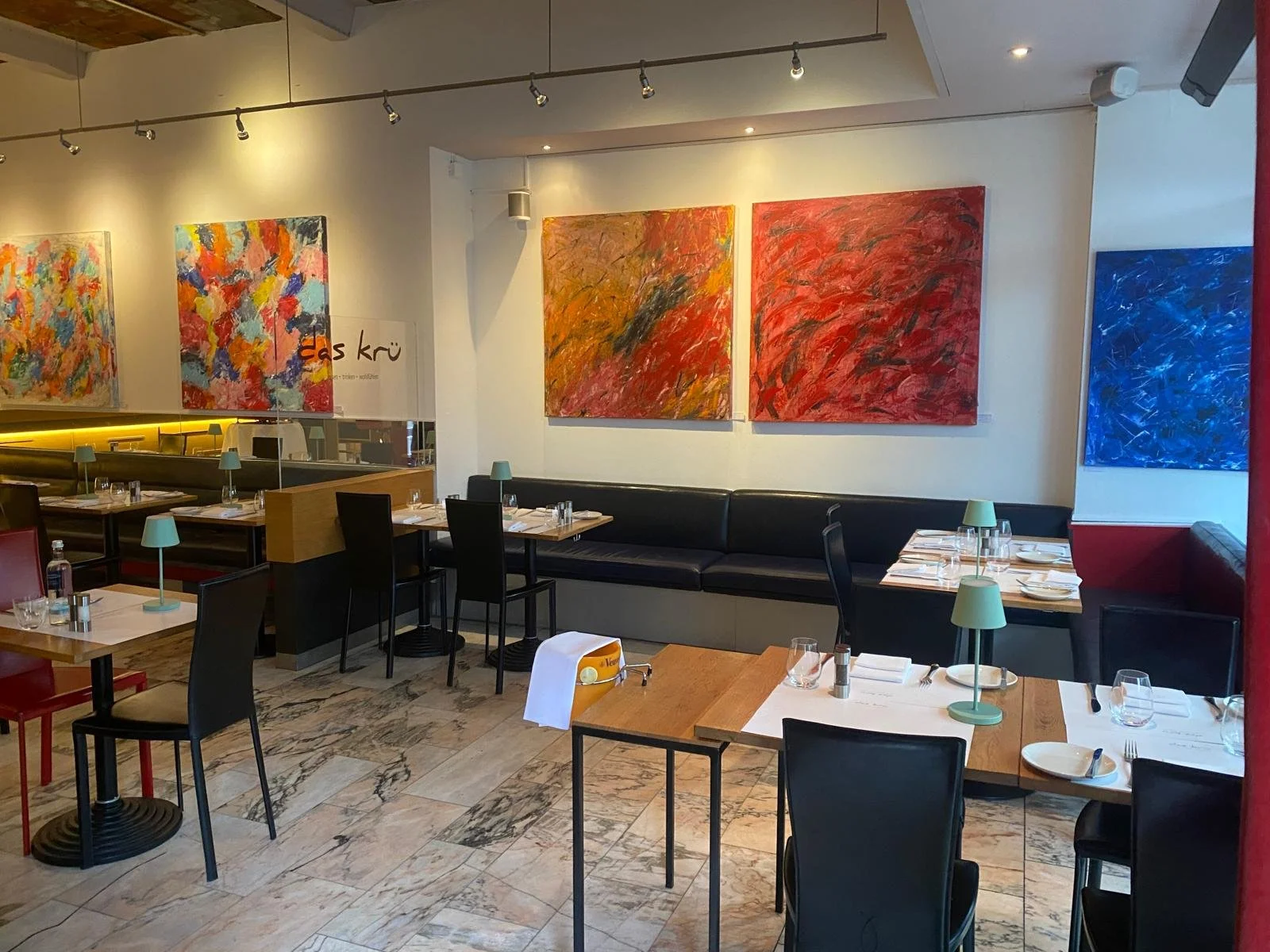 Innenraum eines Restaurants mit mehreren Tischen, schwarzen Stühlen und Wandkunst in bunten abstrakten Farben.