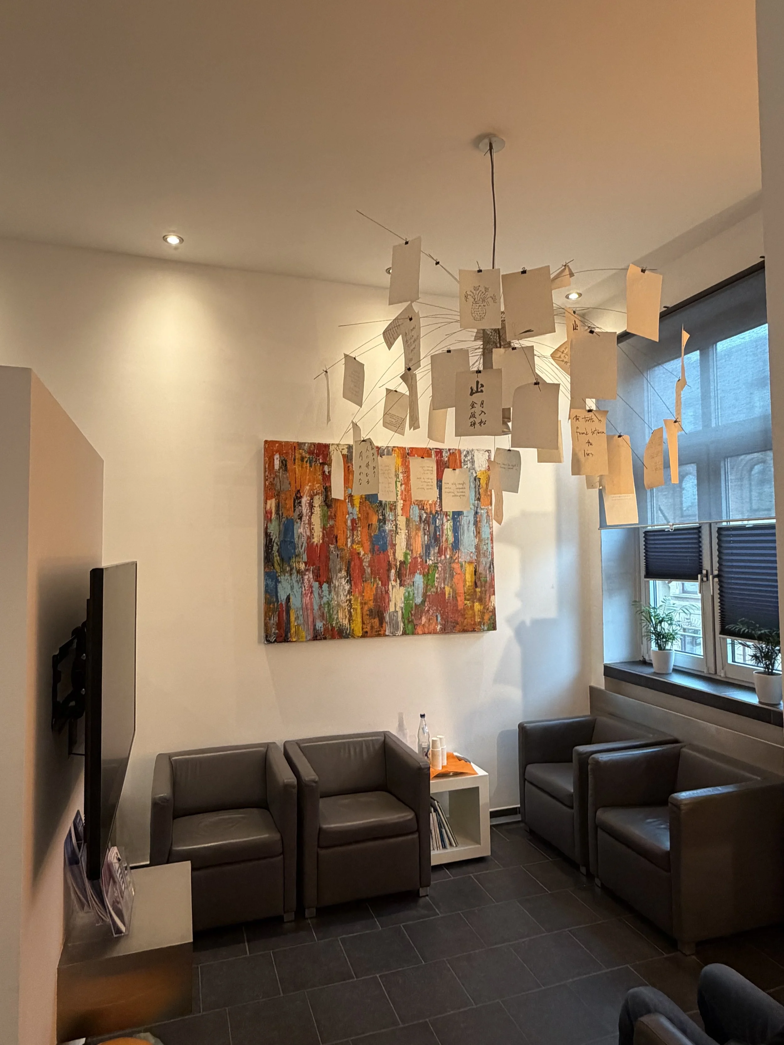 Ein Wohnzimmer mit vier schwarzen Ledersofas, einer weißen Beistelltisch mit orangefarbener Decke und zwei weißen Pflanzen auf dem Fensterbrett. An der Wand hängt ein großes abstraktes Gemälde mit bunten Farben. Die Decke hat ein modernes Lichtdesign, darunter auch eine Lampe mit Karten, die an Schnüren hängen.