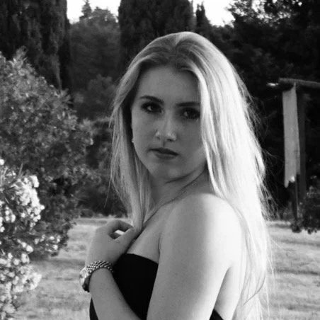 Schwarz-weiß-Bild einer jungen Frau mit langen Haaren, die in einem Garten steht und direkt in die Kamera schaut.
