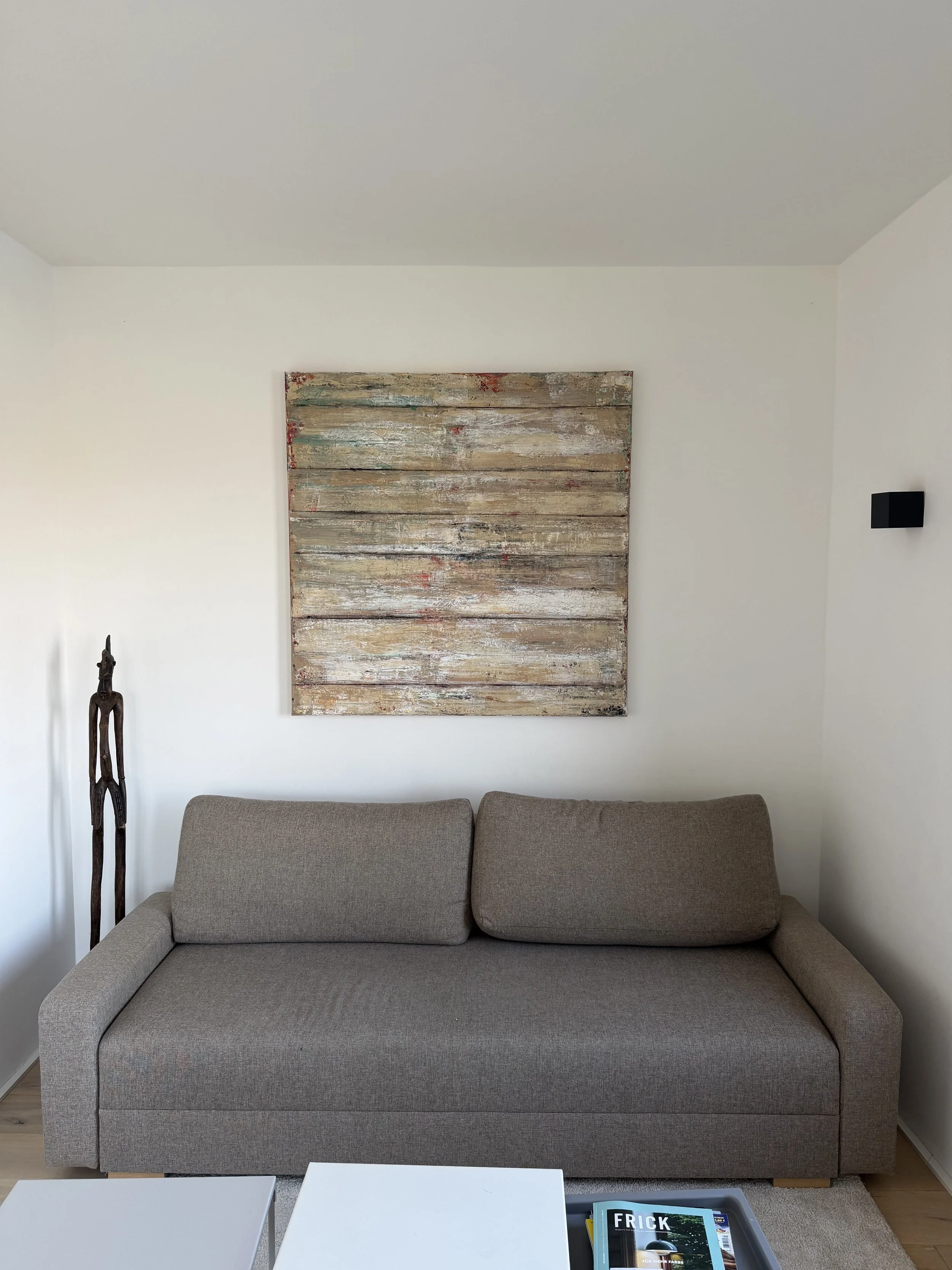 Wohnzimmer mit grauem Sofa, abstraktes Wandbild aus Holz, kleine schwarze Wandlampe, Holzboden, weiße Kaffeetische, Zeitschrift auf Ablage.
