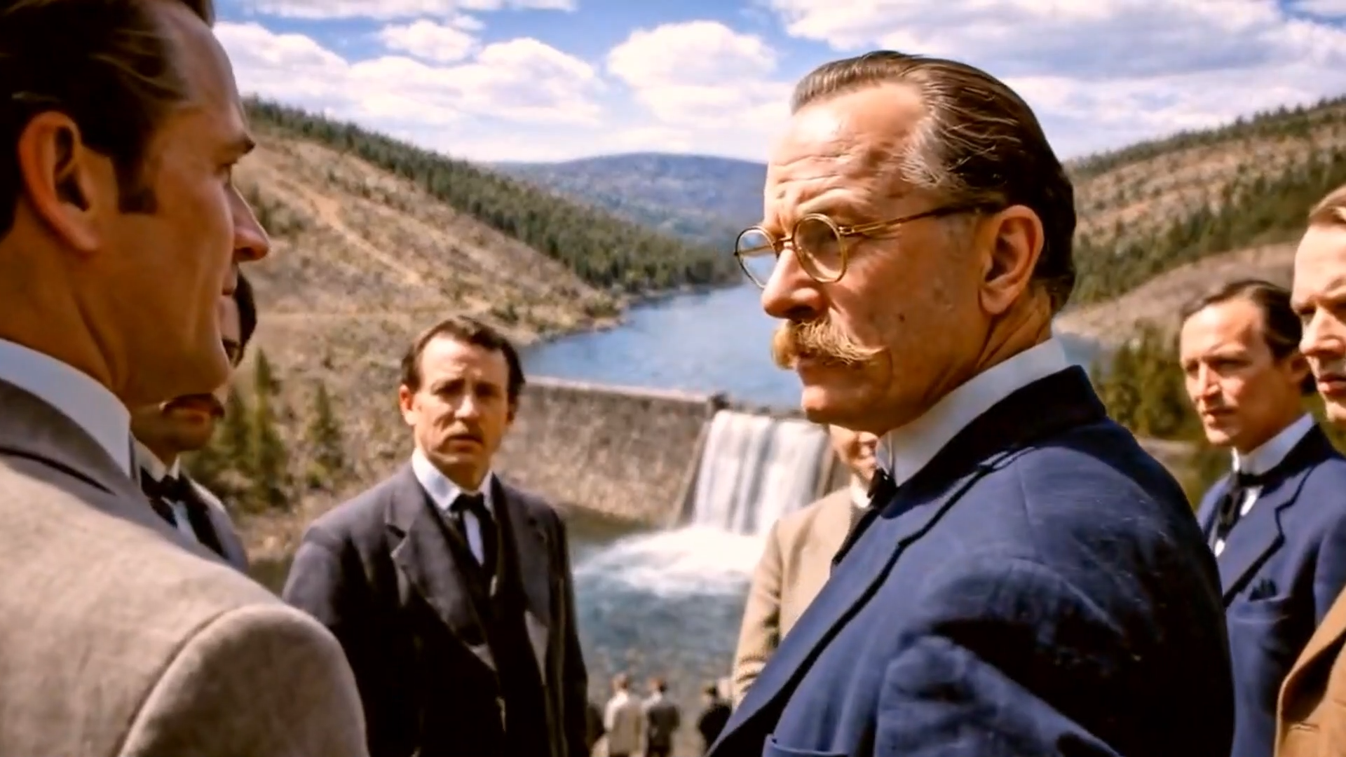 Men in suits and ties talking near a river with a dam and a waterfall in the background, in a hilly, forested area.