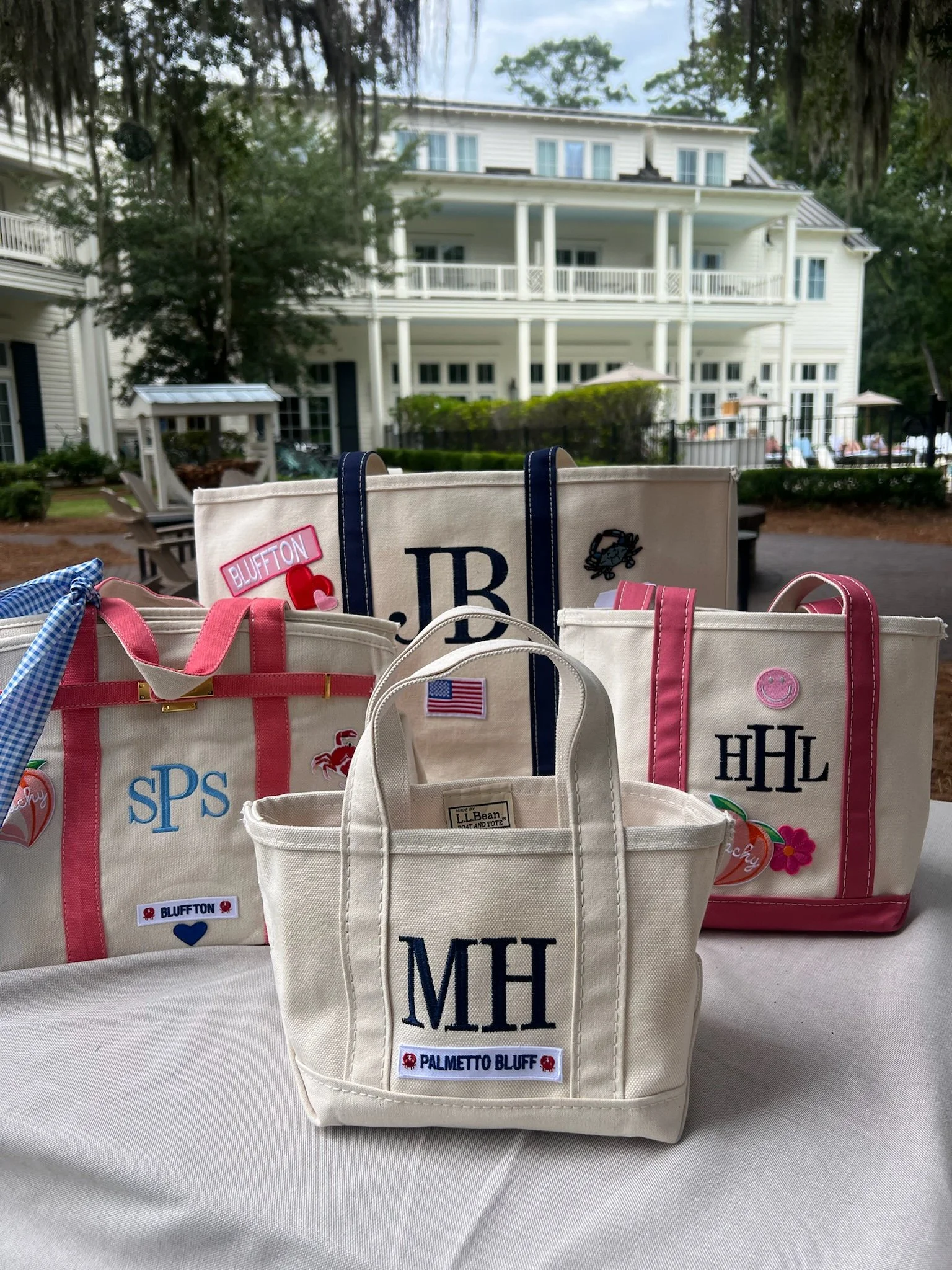 Multiple personalized tote bags with initials and patches, placed on a white table outdoors, with a large white house with balconies and trees in the background.
