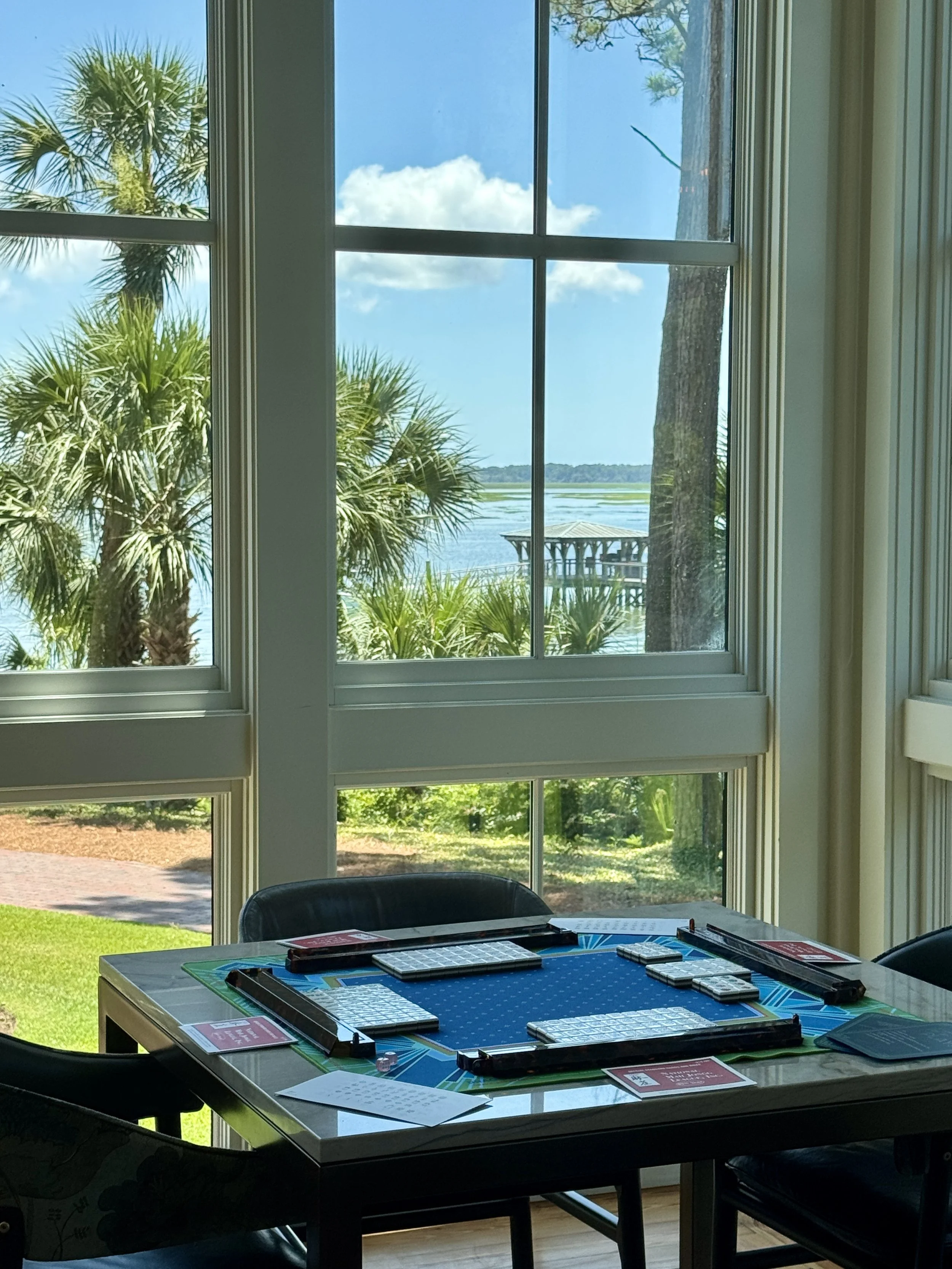 Inside view of a room at the Montage Palmetto Bluff with large windows showing palm trees and a water body outside. A table with mahjong is in the foreground.