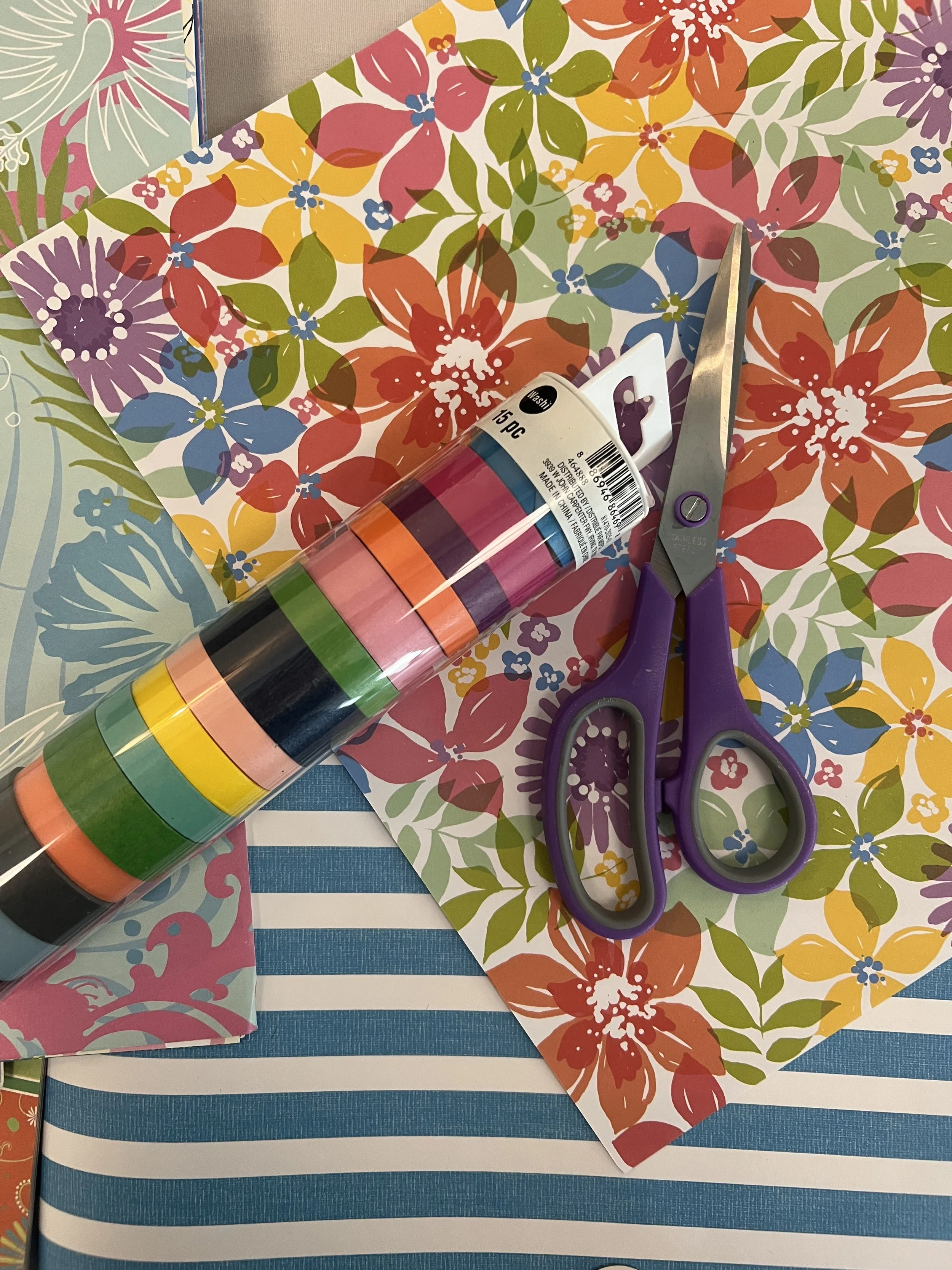 A craft workspace with a colorful floral paper, scissors with purple handles, and a roll of multicolored washi tape on a blue and white striped surface.